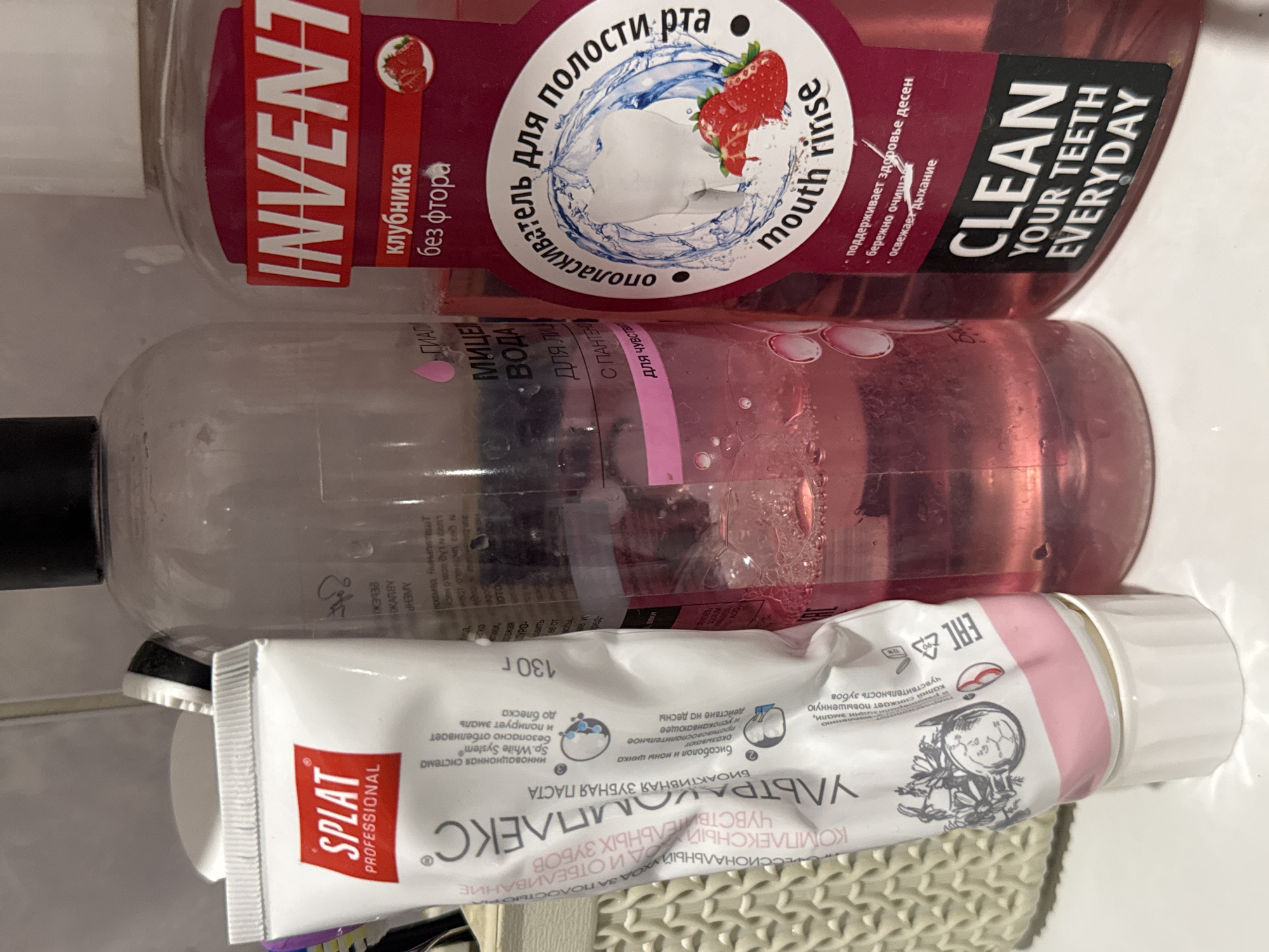 The image shows a collection of oral hygiene products on a bathroom counter. On the left is a tube of SPLAT PROFESSIONAL toothpaste, with pink and white packaging. Next to it is a clear plastic bottle containing pink liquid, likely mouthwash or rinse. To the right is a larger, dark pink bottle with a red label that reads "INVENT" in white text, and further text in Russian indicating "strawberry without fluorine." The bottle also features a label with the words "mouth rinse" in English. The environment appears to be a typical bathroom setting, with a tiled wall visible in the background. The overall impression is one of personal care items arranged for easy access. There are no people or actions depicted in the image.