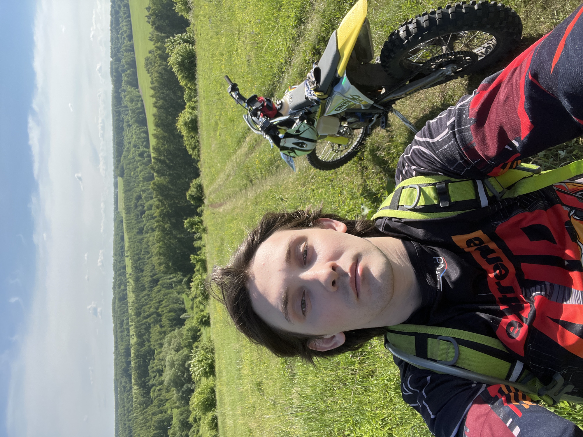 A young adult male takes a selfie on a grassy hill overlooking a vast rural landscape. He has medium-length dark brown hair and a neutral expression, looking directly at the camera. He wears a black long-sleeved motocross jersey with red, orange, and white graphics, along with a bright yellow-green harness. To his right and slightly behind him, a yellow and white dirt bike with knobby tires is parked on a narrow dirt track that ascends the hill. A light-colored helmet and gloves rest on the bike's handlebars and seat. The background features rolling green hills, sparse bushes, and dense forests extending to the horizon under a bright blue sky with scattered white clouds. The scene is set in a rural area, identified as Vorob'evka, Russia.