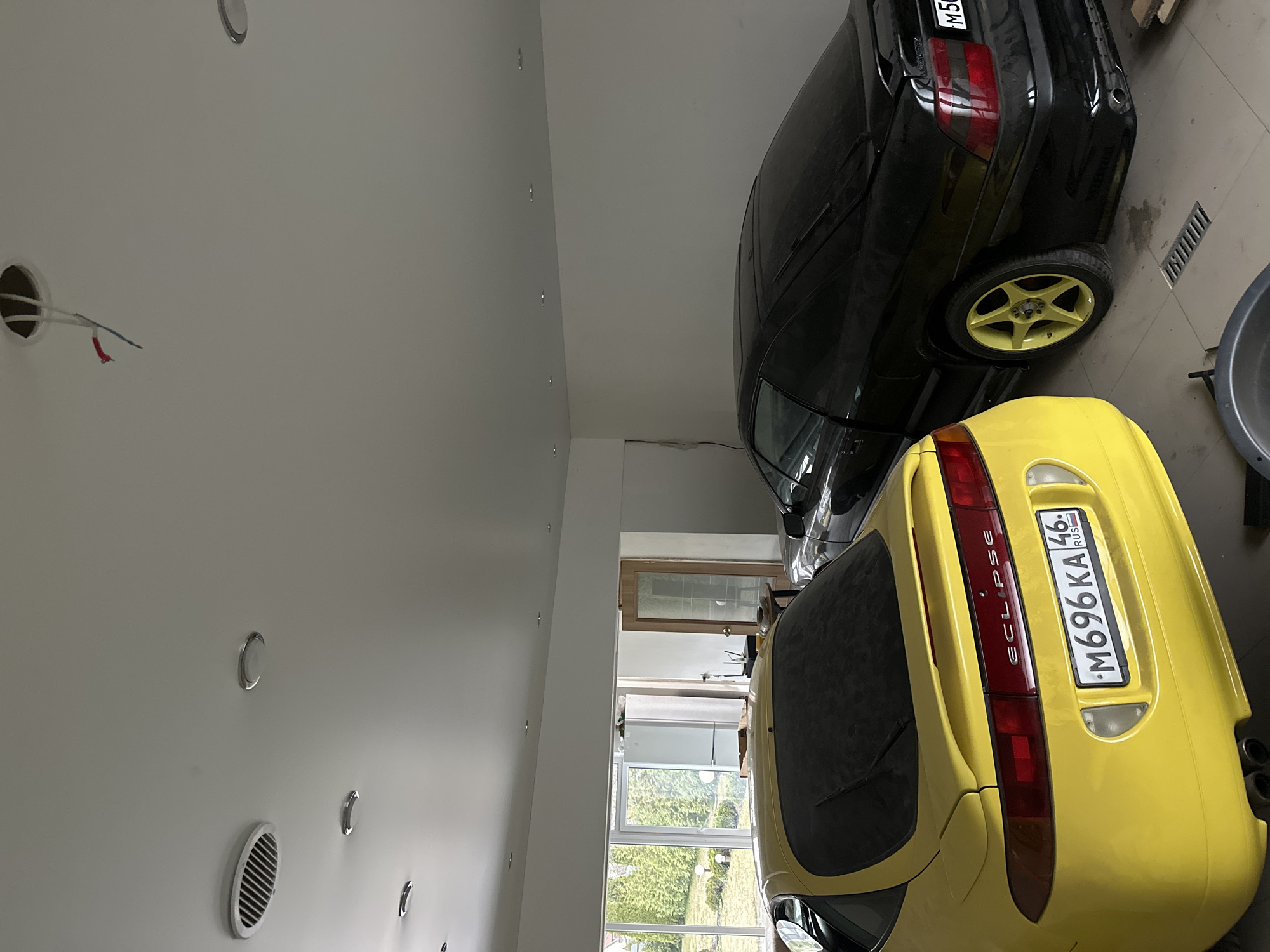 The image shows the interior of a garage. A black car with yellow rims and a yellow car with the license plate "M696KA46" are parked inside. The license plate indicates that the yellow car is registered in the Kursk Oblast region of Russia. The ceiling is white with recessed lighting. A door and windows are visible at the end of the garage.