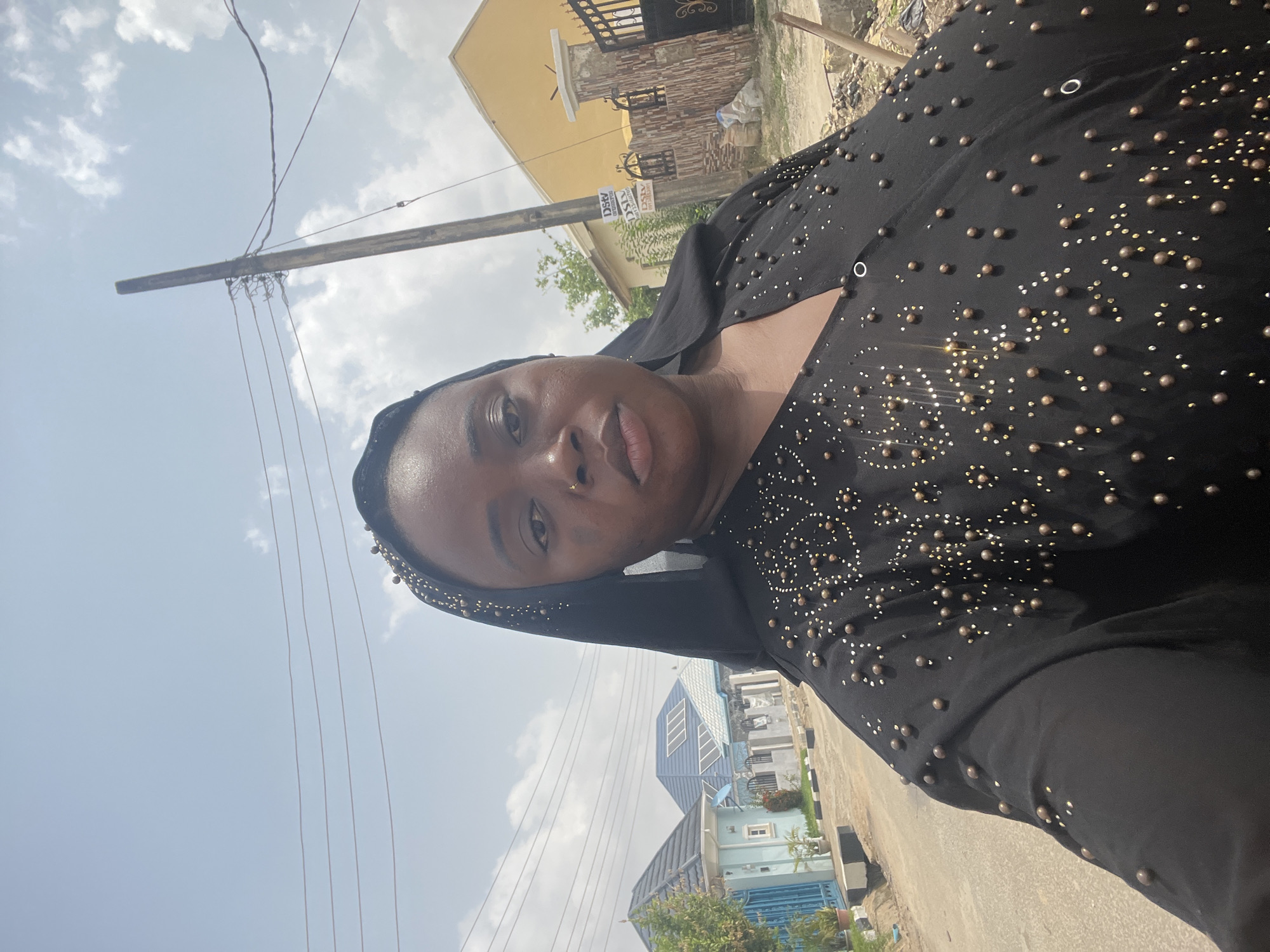 A woman wearing a black, beaded dress and head covering is depicted in a selfie taken outdoors in what appears to be a residential area of Abuja, Nigeria. The sky is partly cloudy. Buildings of various designs and colors are visible in the background. A utility pole with wires runs along the side of the road. The woman is looking directly at the camera with a neutral expression.