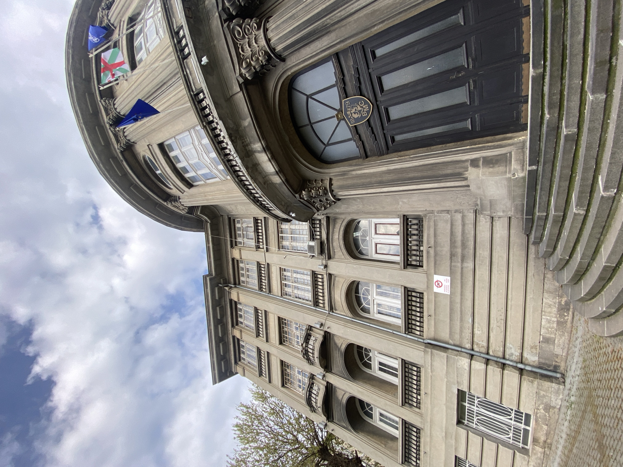 A low-angle view shows the exterior of the Bulgarian Maritime Chamber building in Varna, Bulgaria. The architecture features gray stone with a rounded façade on the left side of the image. Three flags are visible near the top of the rounded structure, with a small sign above a black entrance door. The building has multiple floors with arched windows. A "No Smoking" sign is visible on the building’s exterior. The sky with scattered clouds fills the space on the lower left corner.