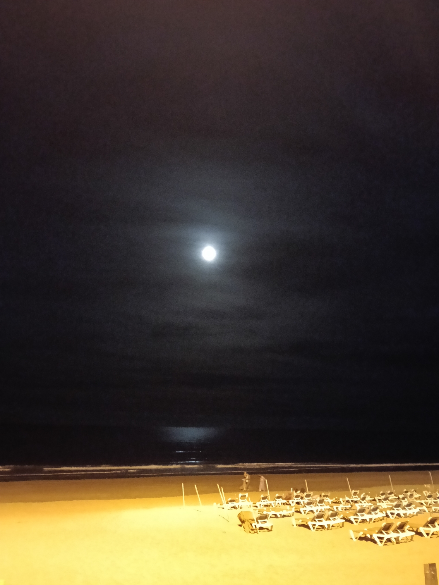 Here is a forensic description of the image:
The image depicts a nighttime scene on a beach in San Bartolomé de Tirajana, Spain. The sky is dark with a full moon prominently visible. Its reflection appears on the ocean water.
A sandy beach occupies the lower portion of the image, with rows of arranged beach chairs. In the midground of the beach, two figures stand together near the water’s edge. The horizon line is visible, separating the ocean from the sky.