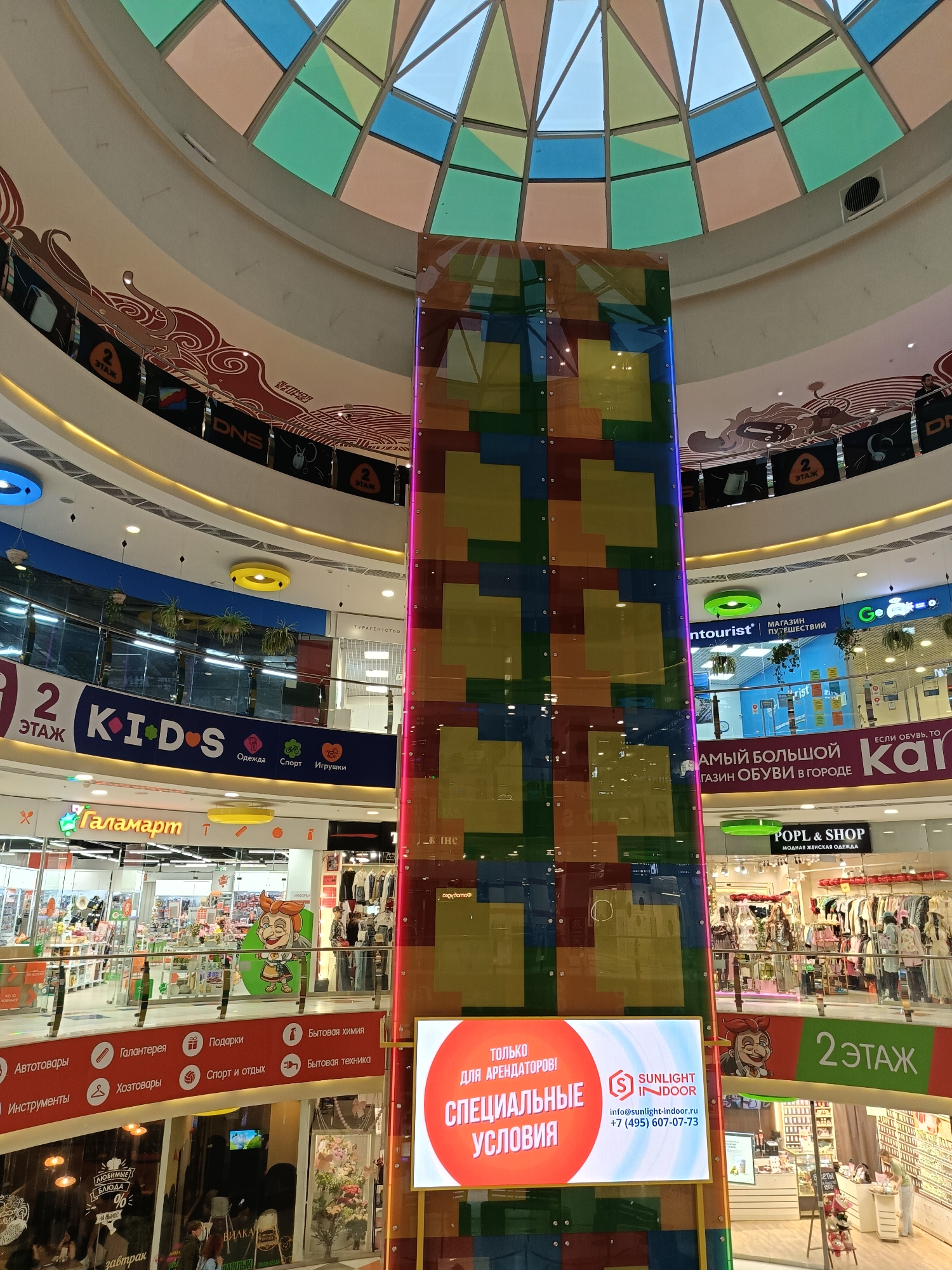 This is an indoor shot of the interior of a shopping mall in Saint Petersburg, Russia. The atrium rises several floors, capped with a colorful geometric dome letting in natural light. A tall, square column covered with multicolored squares runs vertically through the center of the frame, with a large advertisement in Russian stating "ONLY FOR TENANTS! SPECIAL CONDITIONS" from Sunlight Indoor. The mall features various stores on multiple levels, including "KIDS" with clothing, sports and toy sections, "Галамарт" (Galamart), "ntourist," "Kai," and "POPL & SHOP," catering to different shopping needs. Each floor is marked with a large "2 ЭТАЖ" sign, indicating the second floor in Russian. People are visible browsing the stores on the lower levels. The mall incorporates plant decorations hanging from ceilings and railings, adding to the overall ambiance.