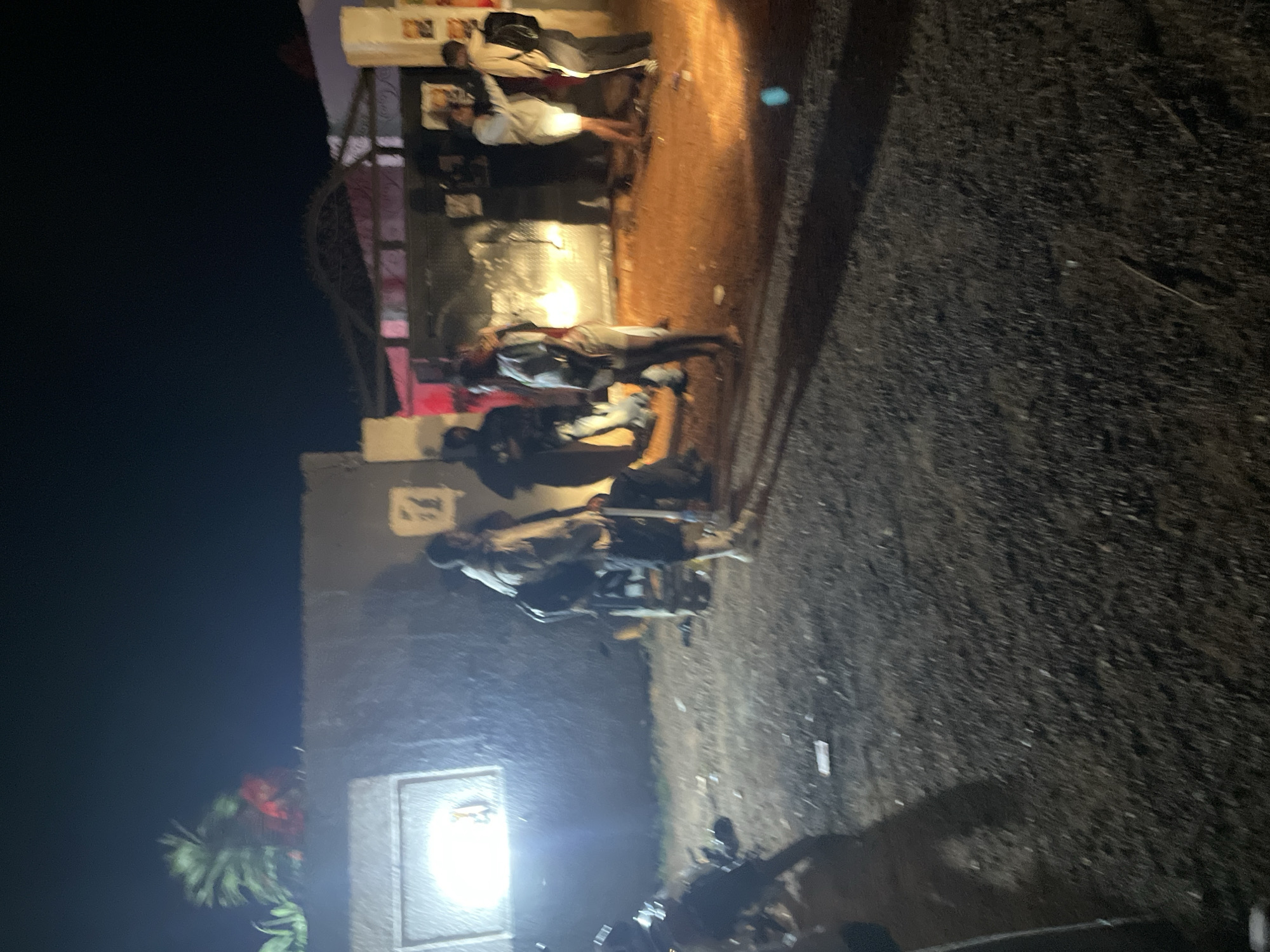The image depicts an outdoor scene at night, likely in Durumi, Nigeria, characterized by a dimly lit environment with several individuals present. A group of people, appearing to be young adults, are gathered near a building with an open gate. The building's facade is dark, with a bright light emanating from a window on the left side of the image. A large, ornate gate with an arched top is visible in the center, leading into the property.
The ground is covered with gravel and dirt, with some scattered debris visible. Some of the individuals are carrying backpacks. The lighting suggests artificial illumination, possibly from the building or temporary lights set up for an event. The overall atmosphere appears to be one of a gathering or entrance to a venue at night.