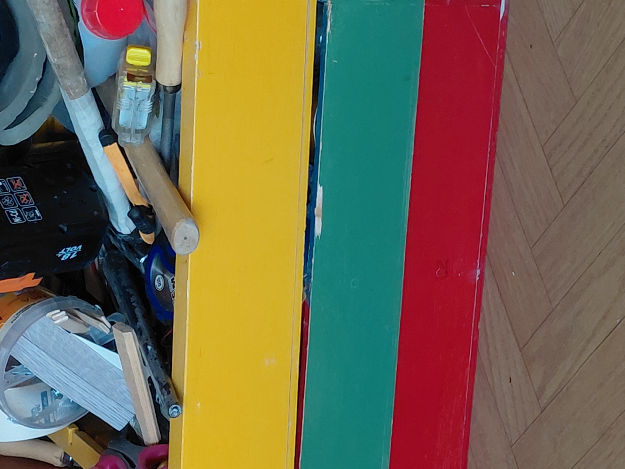The image captures a detailed view of a cluttered indoor space, likely a storage area or workshop, with a distinctive background. The left side of the frame is dominated by an assortment of items, including various tools and components. A red bottle cap is visible near the top, alongside a clear plastic object with a yellow cap, possibly a small bottle or glue stick. Several wooden handles of tools are interspersed throughout the clutter. A prominent black object features white text clearly reading "18 VOLT" along with several warning icons depicted as crossed-out squares, suggesting it is a battery pack or part of a cordless tool. Other items include a clear cylindrical container filled with small wooden sticks, a stack of thin white rectangular pieces, parts of an orange tool, and red-handled scissors.
To the right, three distinct, brightly painted vertical boards create a colorful background or partition. From left to right, these boards are yellow, green, and red. They appear to be made of wood or a similar material, showing some wear and slight gaps between them. The very bottom right of the image reveals a wooden floor laid in a herringbone pattern, indicating an indoor setting. No people are visible, and no specific activity is taking place beyond the items being stored. The lighting suggests an indoor environment, but the time of day is not discernible. A faint, small "R" or similar mark is visible on the red board near the top.