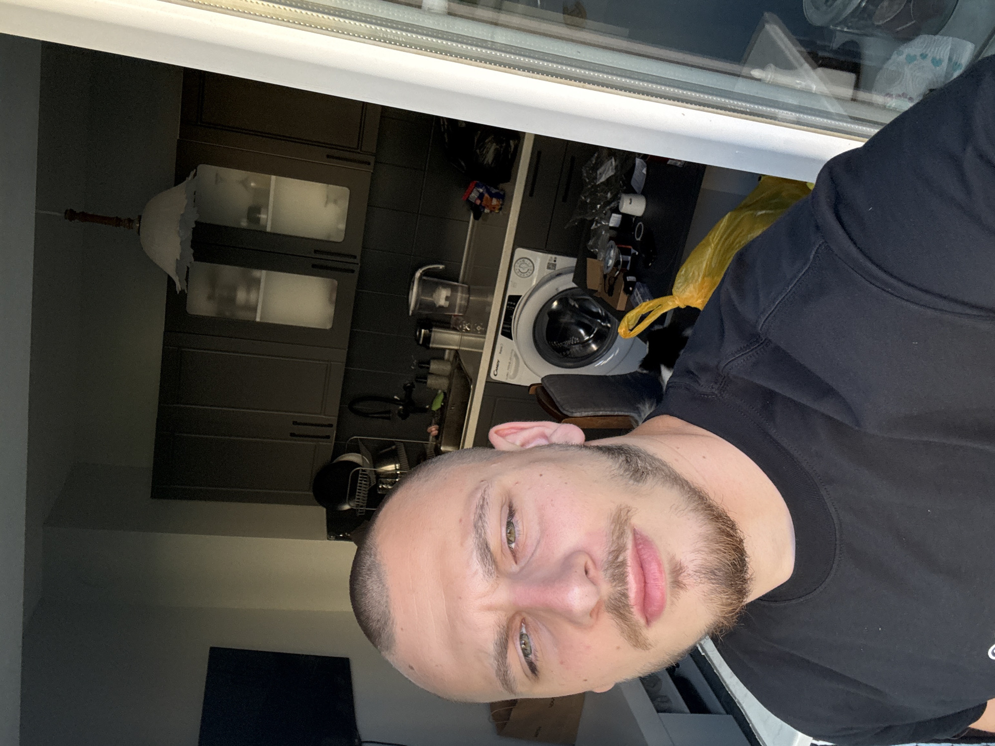 The image shows a close-up selfie of a young man with a buzz cut and a beard, wearing a black t-shirt. He is indoors, in what appears to be a kitchen or utility area. Behind him, a washing machine is visible, along with kitchen cabinets, a sink, and various household items. The lighting suggests it is daytime, possibly early morning or late afternoon, given the shadows. The overall setting is cluttered and informal. The man's expression is neutral. The location is identified as Stara Zagora, Bulgaria.