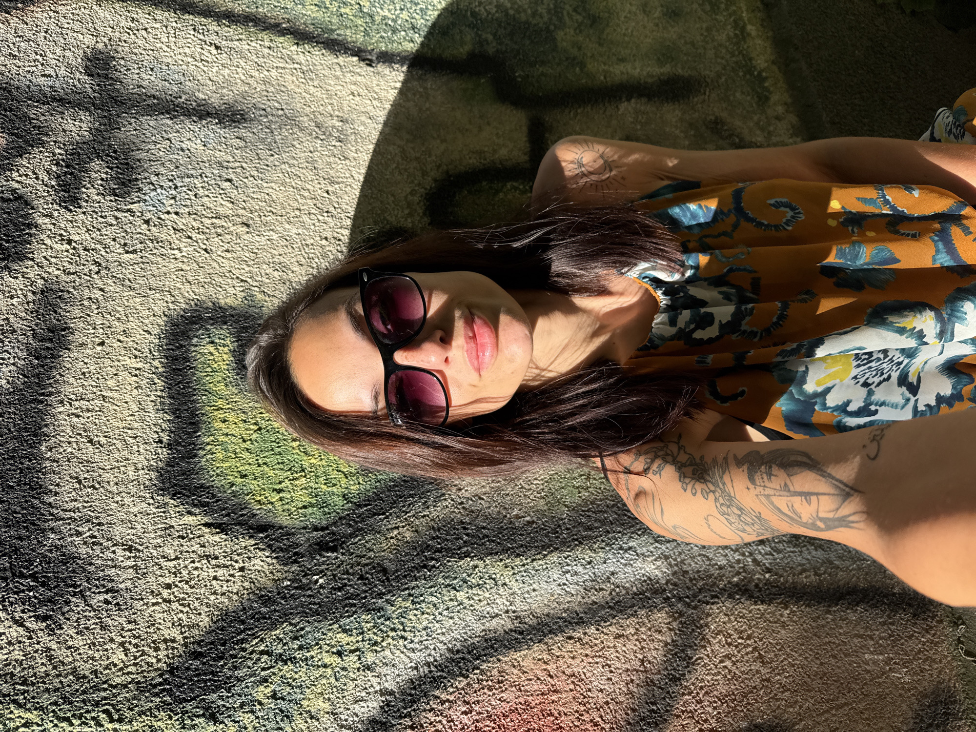 A woman wearing sunglasses and a floral print top is lying on a textured, painted surface. The painting shows layers of colors including beige, green, black, and traces of red. The woman has dark hair and light skin. Her arms are visible, and she has tattoos on them. The scene is lit with dappled sunlight creating sharp shadows on the surface where she is lying. The location in the city of Pleven, Bulgaria is unknown.