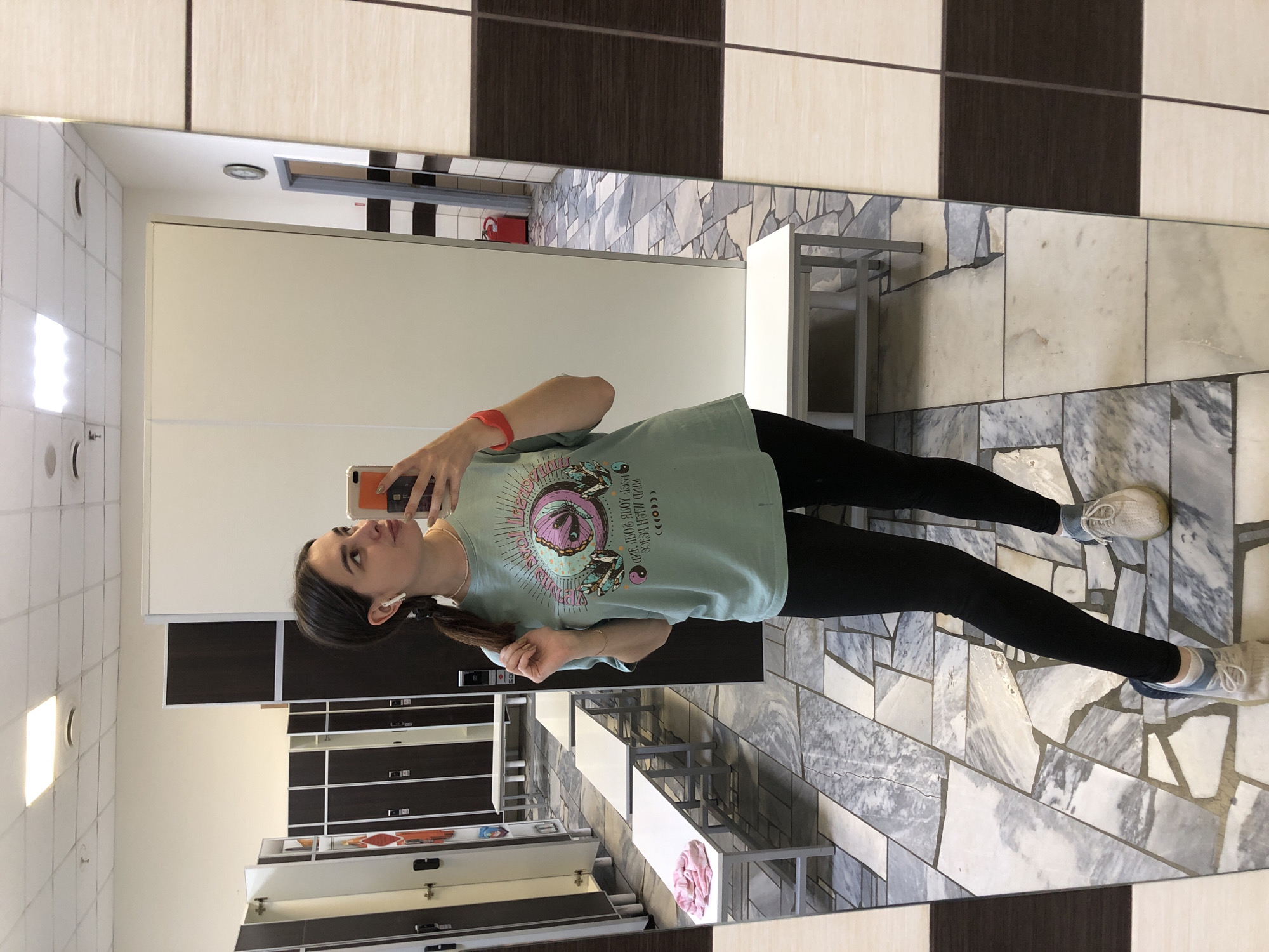 A young woman, approximately 20-25 years old, is taking a mirror selfie in what appears to be a locker room. She has dark brown hair pulled back in a ponytail and is wearing a light blue t-shirt with a graphic design, black leggings, and athletic shoes. She has wireless earbuds in and a red fitness tracker on her left wrist. The locker room has rows of metal lockers, white benches, and a tiled floor. In the background, a fire extinguisher is visible. The overall impression is that of a gym or sports facility. The image is likely taken in the city of Chaikovsky, Russia, based on the provided location information.