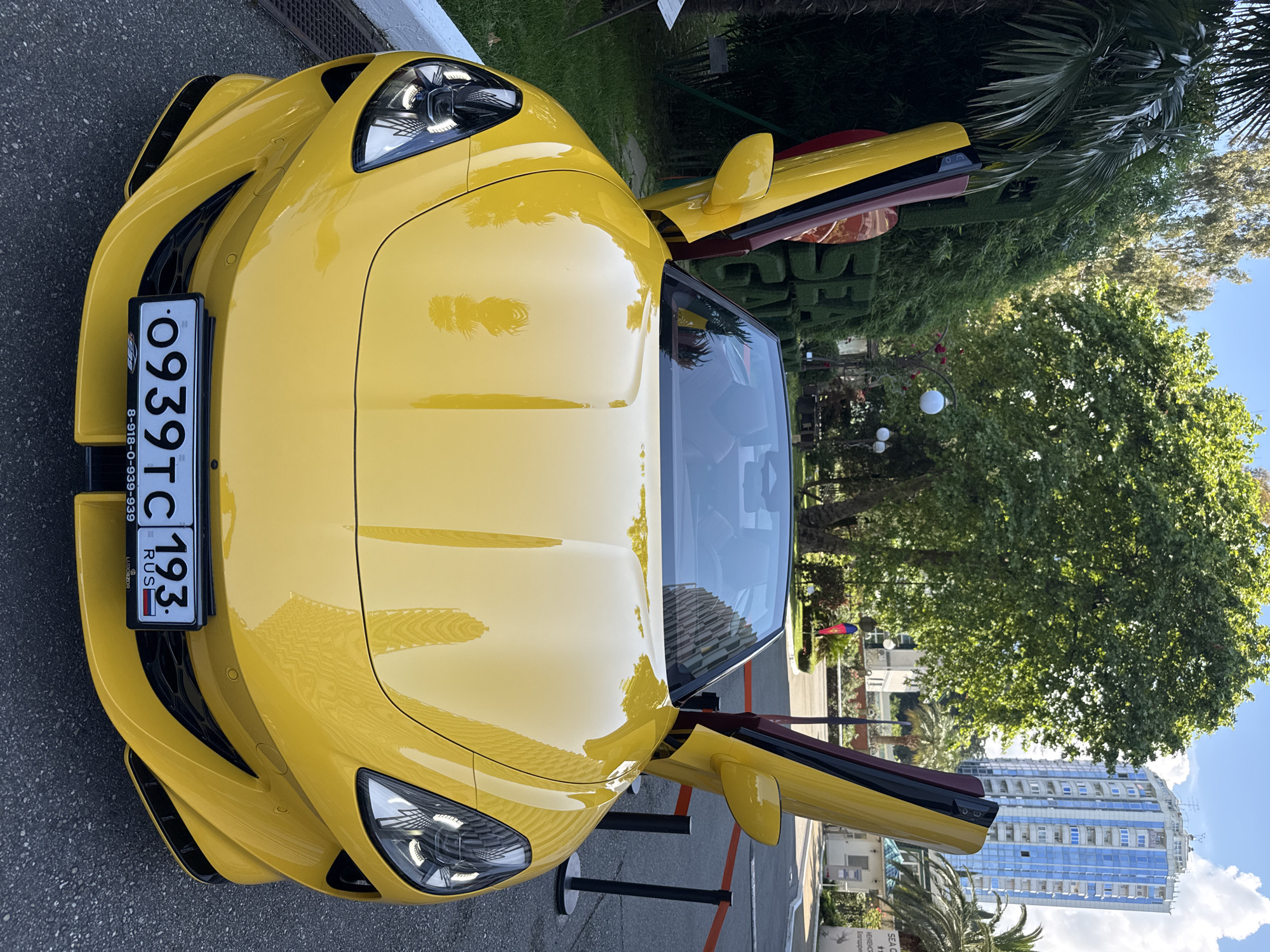 A bright yellow convertible Ferrari with its doors open is parked on an asphalt surface. The car faces forward, with its headlights on. The license plate reads "0939TC 193 RUS". In the background, there is a hedge sculpture spelling out "SEA GARDEN", a tall blue building, palm trees, and lush green trees under a bright sky. The scene appears to be in Sochi, Russia, based on the building architecture and vegetation.