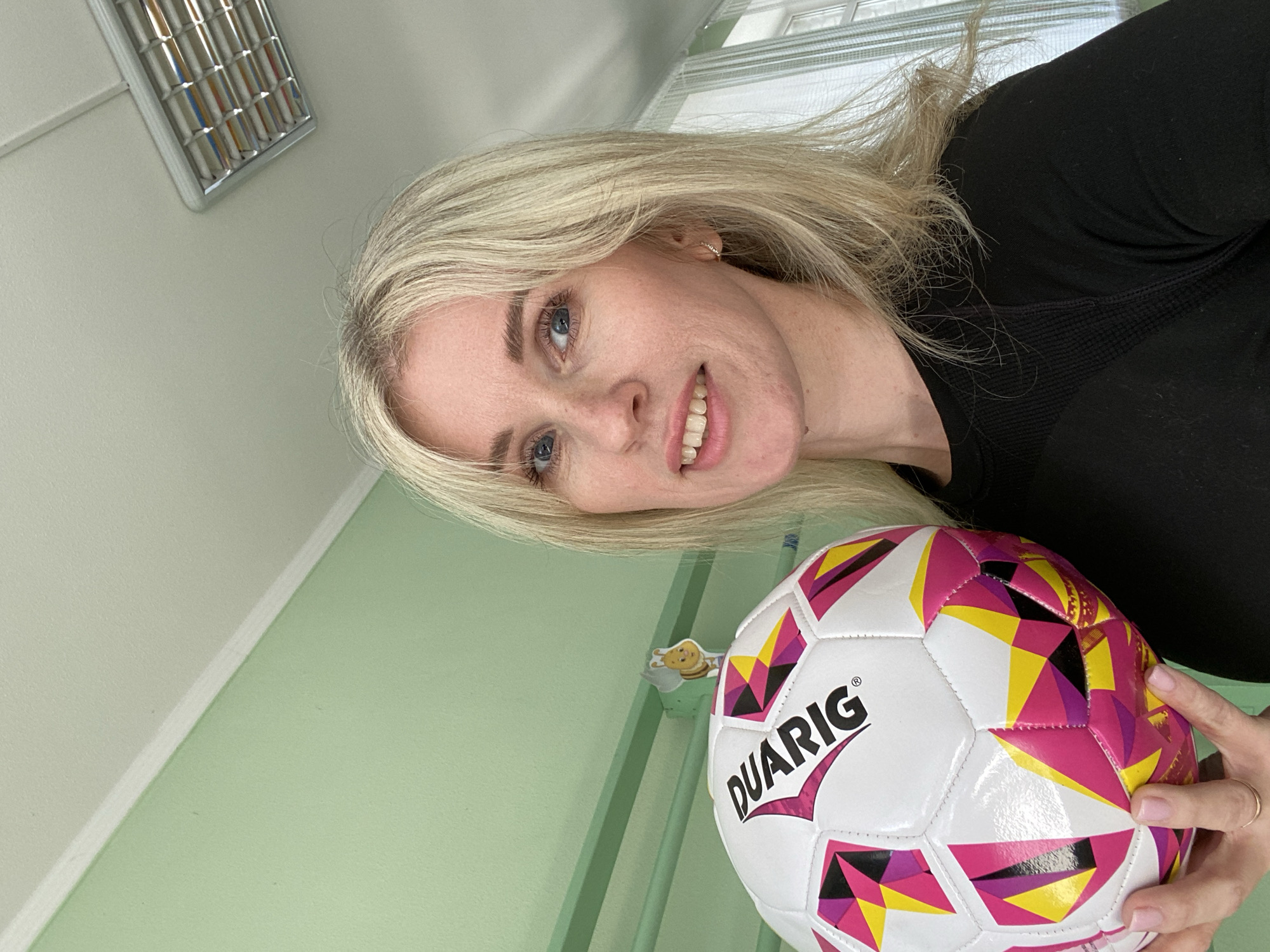 A woman with blonde hair is holding a colorful soccer ball with the word "DUARIG" printed on it. She is wearing a black top and is smiling. The background is a light green wall with a metal vent on the ceiling. The location is an interior space. The city and country are unknown.