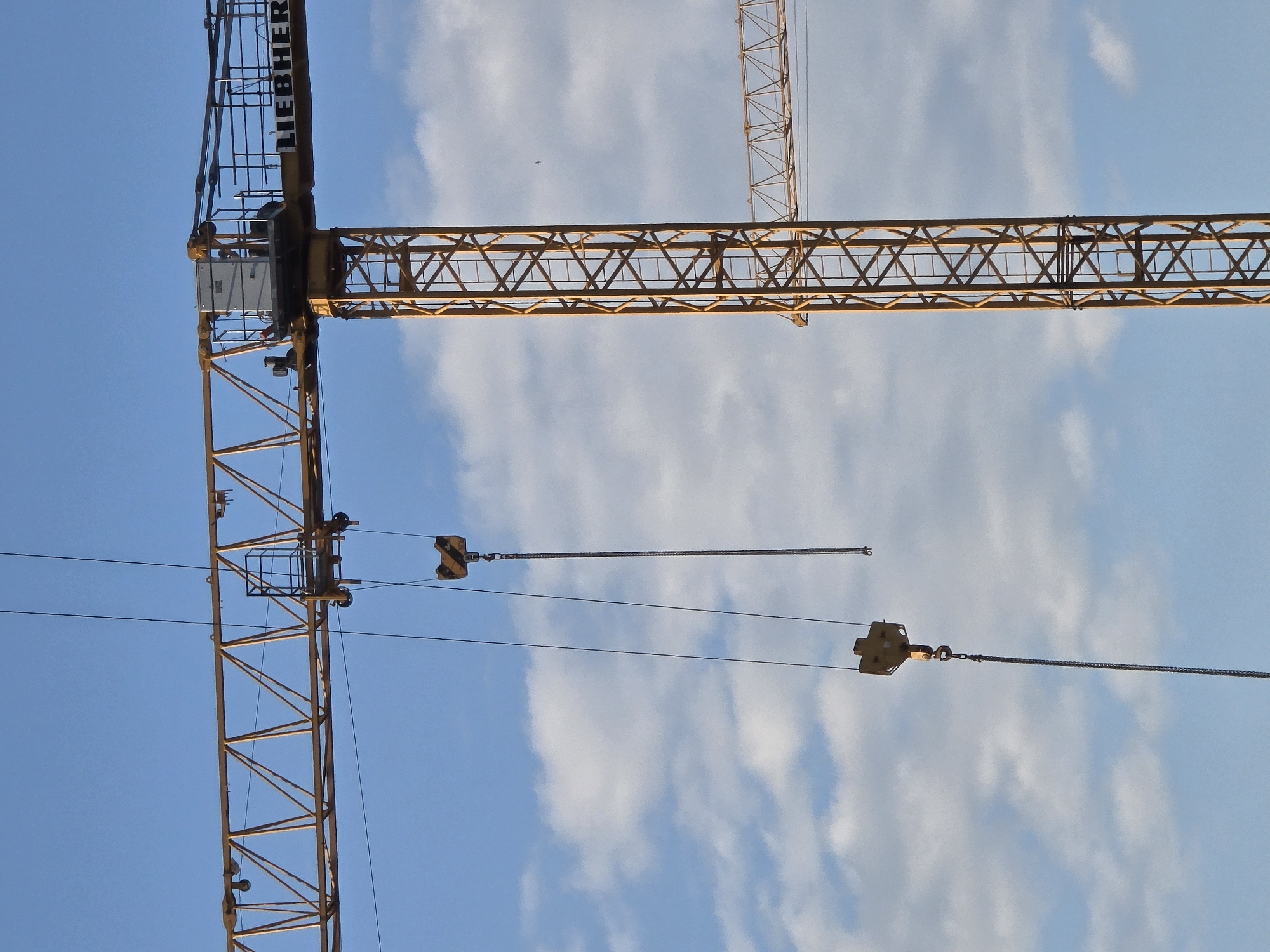 The image captures a Liebherr crane against a partly cloudy blue sky in Unknown, Unknown. The crane is constructed of orange steel and consists of a vertical tower, a horizontal boom, and a cable. The brand "Liebherr" is visible on the vertical support tower. The cable extends from the boom and has two orange blocks that look like pulley systems at each end.