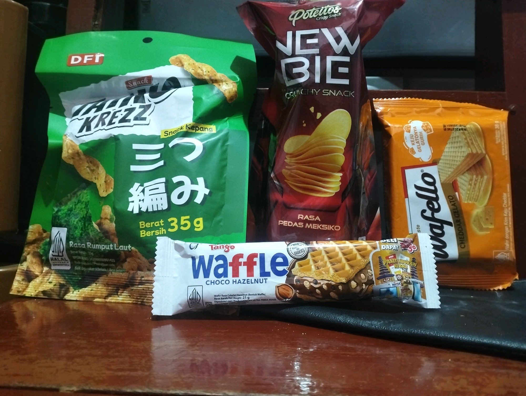 In a low-angle shot, several snack food packages are arranged on a dark brown, reflective surface, possibly a table. The background is dark and out of focus. From left to right, the packages include: a bright green bag with the "DFI KREZZ" logo and Asian characters, suggesting a seaweed or vegetable snack. Next to it is a tall, maroon bag for "Poteitos NEW BIE Crunchy Snack," featuring images of potato chips. To the right of that is an orange bag labeled "wafer" with a picture of wafer crackers, and the text "KEJU GELATONYA GURIH." In the foreground, a white and blue wrapper for "Tango waffle CHOCo HAZELNUT" is prominently displayed, showing a waffle bar with chocolate and hazelnut. The overall scene is a still life of packaged snacks. The specific location of Klecorejo, Indonesia, cannot be determined from the provided image.