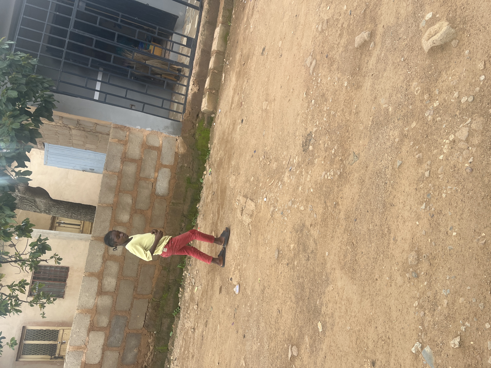 A young girl with braided hair, wearing a yellow t-shirt and red pants, walks on a dirt path in front of a brick wall. She is wearing dark sandals. Behind her, a building with barred windows and a metal gate is visible. The ground is covered with dirt, small rocks, and scattered debris. Trees with green leaves hang over the scene from the top left. The location is Gura Top, Nigeria.