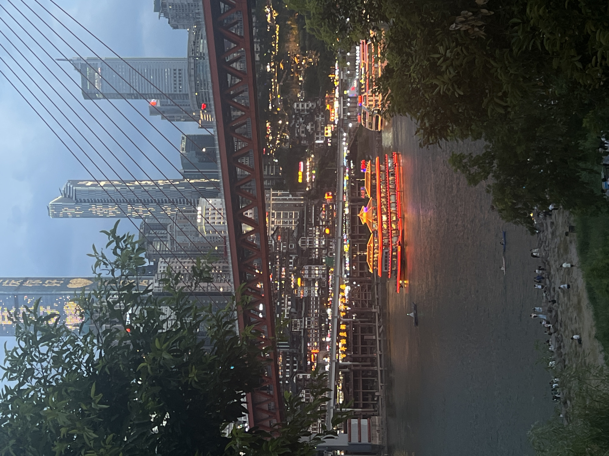 The image depicts a twilight scene in Jiangbeicheng Street, China, with the Yangtze River flowing in the foreground. Several illuminated boats are on the river, with a prominent multi-tiered passenger boat featuring bright red and yellow lights in the center. The riverbank is lined with people gathered on rocks and grass. Across the river, a dense urban landscape rises, characterized by numerous buildings with illuminated windows, creating a vibrant cityscape. A large, red suspension bridge spans the river, its structural details visible against the dusky sky. In the background, modern skyscrapers with patterned lights are prominent. Trees with dense foliage frame the left and right sides of the image, obscuring parts of the cityscape and adding depth to the scene. The sky is overcast and dark, suggesting either dusk or dawn.
