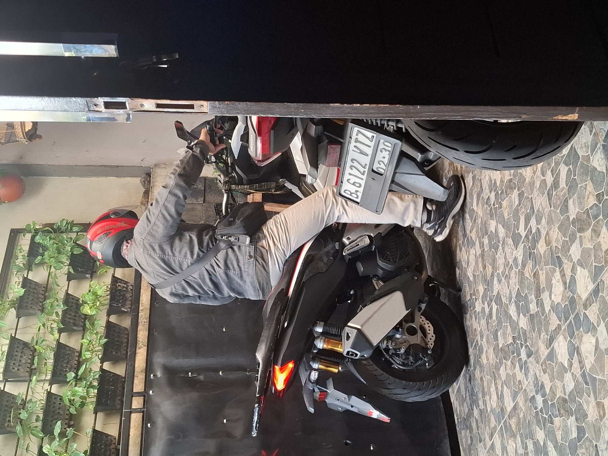 A person wearing a red and black helmet and a gray jacket is sitting on a black scooter. The scooter has a license plate that reads "B. 6122 VTZ" and "02-30". The scooter is parked on a tiled floor. In the background, there is a wall with plants in planters. The scene appears to be in a residential or commercial area. There is no specific indication of this being in West Jakarta, Indonesia.