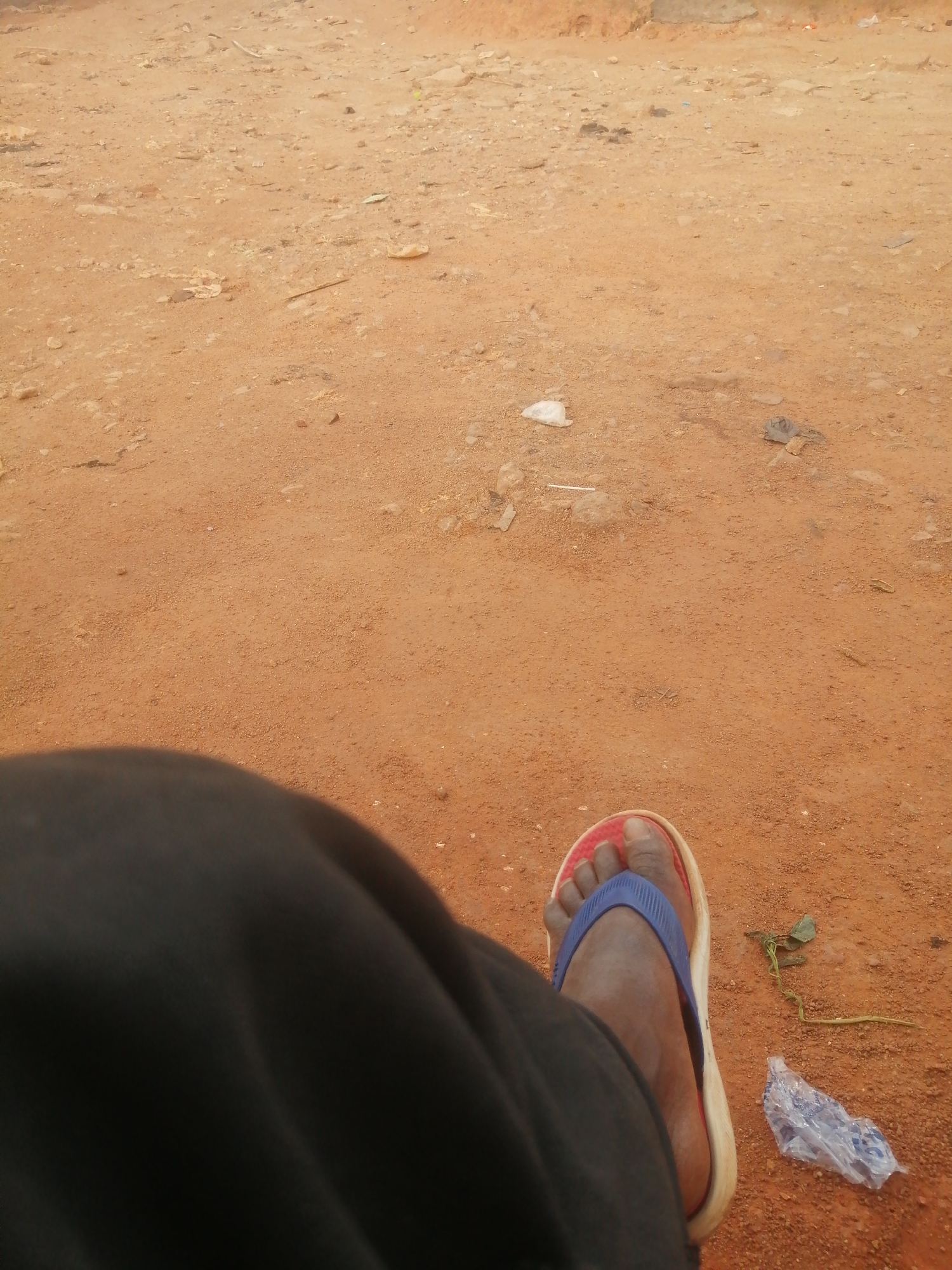 The image shows a close-up view of a person's right foot wearing a flip-flop sandal, resting on a dirt ground. The ground is reddish-brown and appears to be a natural surface, possibly in a rural or less developed urban area. There is some scattered debris on the ground, including small pieces of plastic and a dried leaf. The person is wearing dark trousers that cover most of their leg. The scene is lit by natural daylight. The image appears to have been taken in Jos, Nigeria, given the typical soil color and landscape often found in the region.