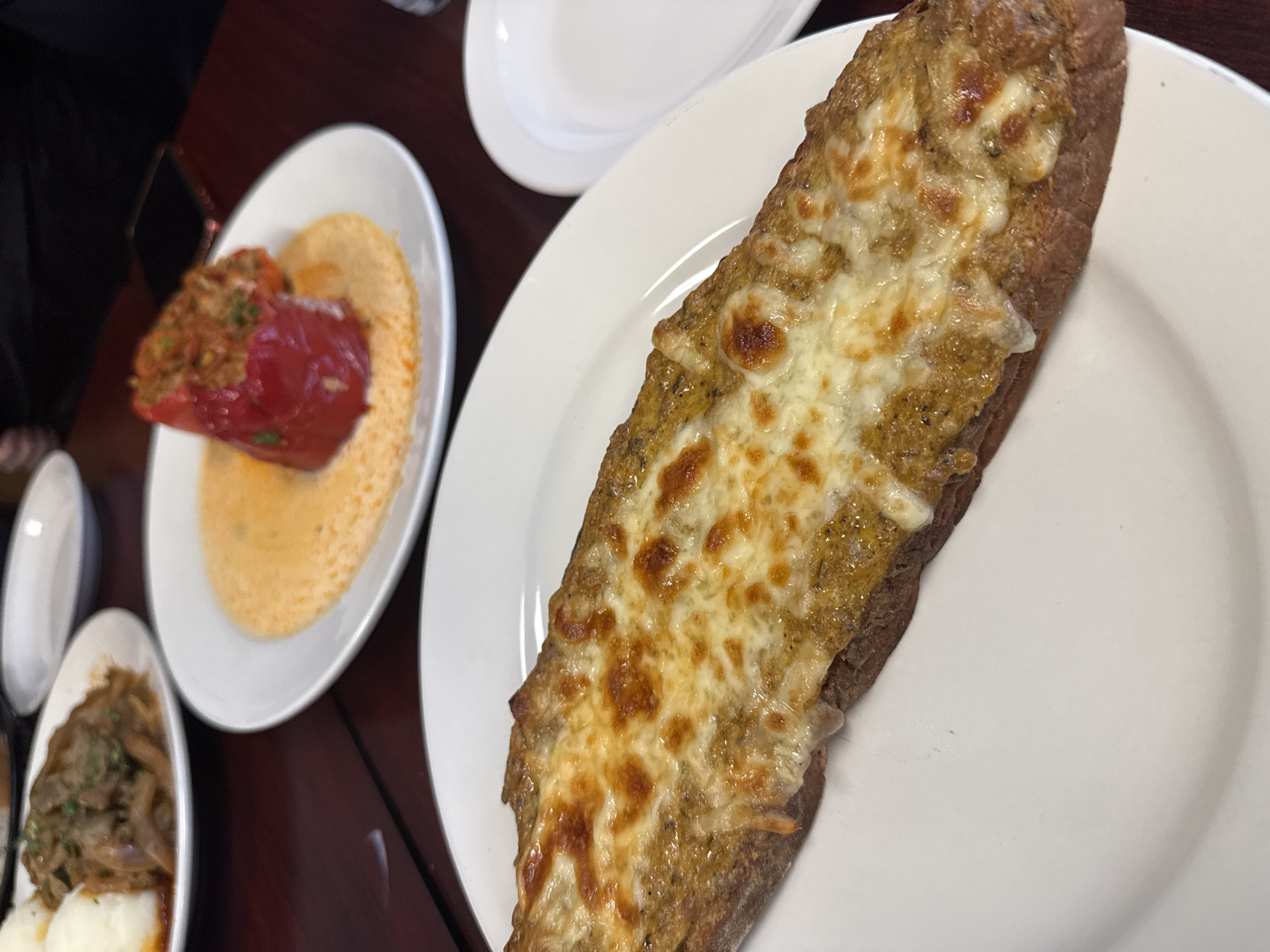 The image shows a table setting with three white plates on a dark wooden surface. On the right is a white plate with a baguette topped with melted cheese. The bread has a golden-brown crust and the cheese is bubbly and browned in places. In the center is another white plate with a stuffed red bell pepper atop a yellow puree. To the left is a smaller white plate with a portion of mushrooms and what appears to be mashed potatoes. A small white dish is also visible to the left. A portion of a person's dark clothing is visible in the upper left corner. Based on the presence of the stuffed pepper, the location may be the Allegretti's Bakery Cafe in Des Plaines, Illinois.