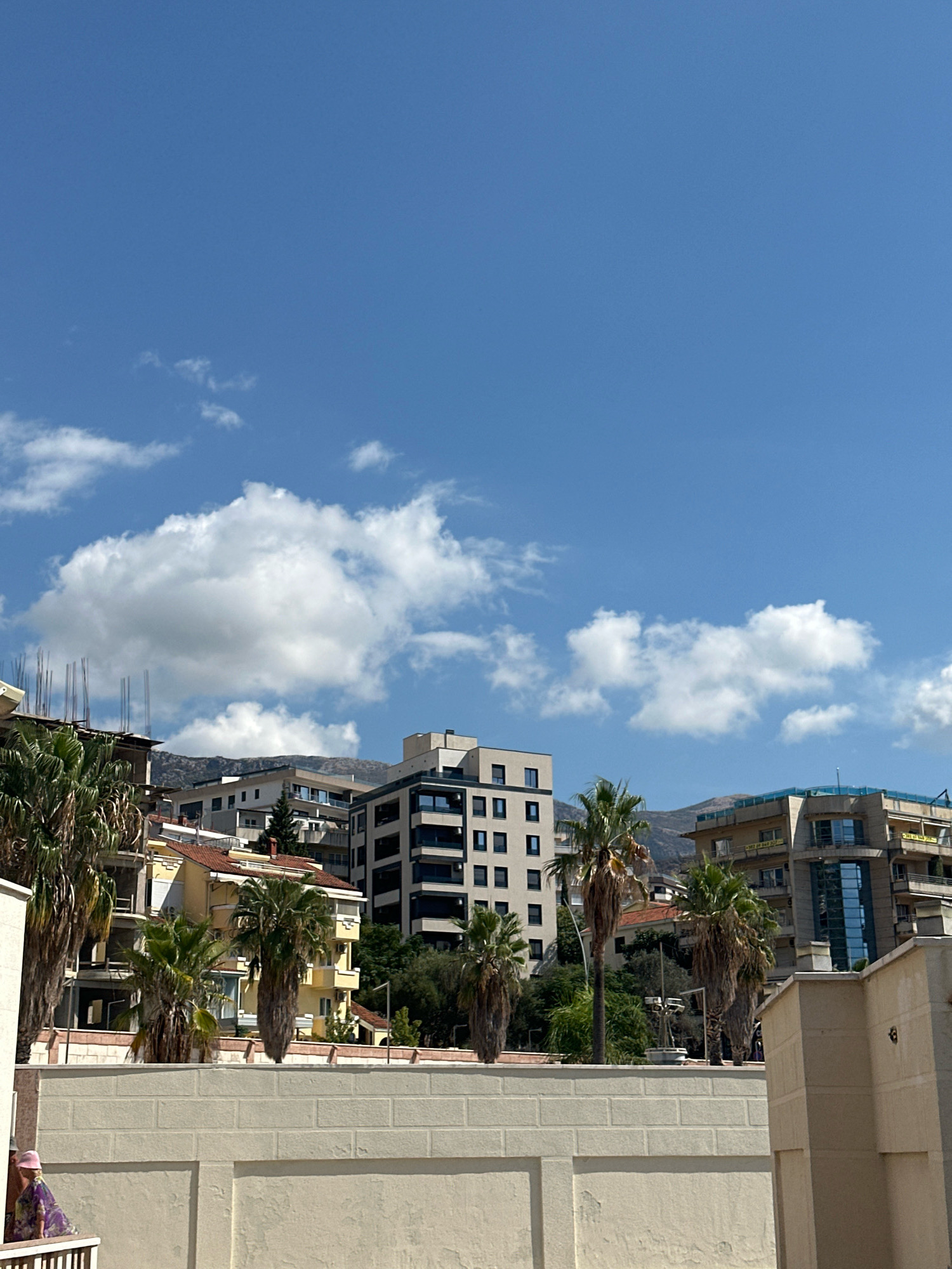 The image shows a view of Boreti, Montenegro, on a clear day. In the foreground, a cream-colored brick wall runs horizontally across the lower part of the frame, with some signs of wear and tear. To the left, a person wearing a pink hat and a patterned dress stands partially visible on a balcony, looking out at the scene. Behind the wall, a line of buildings rises, interspersed with palm trees.
The buildings vary in architectural style and age. Some appear to be under construction, with exposed concrete and rebar. Others are completed, featuring a mix of modern designs with large glass windows and older structures with red-tiled roofs. Among the completed buildings is a taller, contemporary structure with a dark facade and multiple floors. Further in the background, a range of hills or mountains forms a natural backdrop.
The sky above is a vibrant blue, with scattered white, puffy clouds. The overall impression is of a sunny, urban environment, possibly a coastal town given the palm trees and the clear skies often associated with such locations.