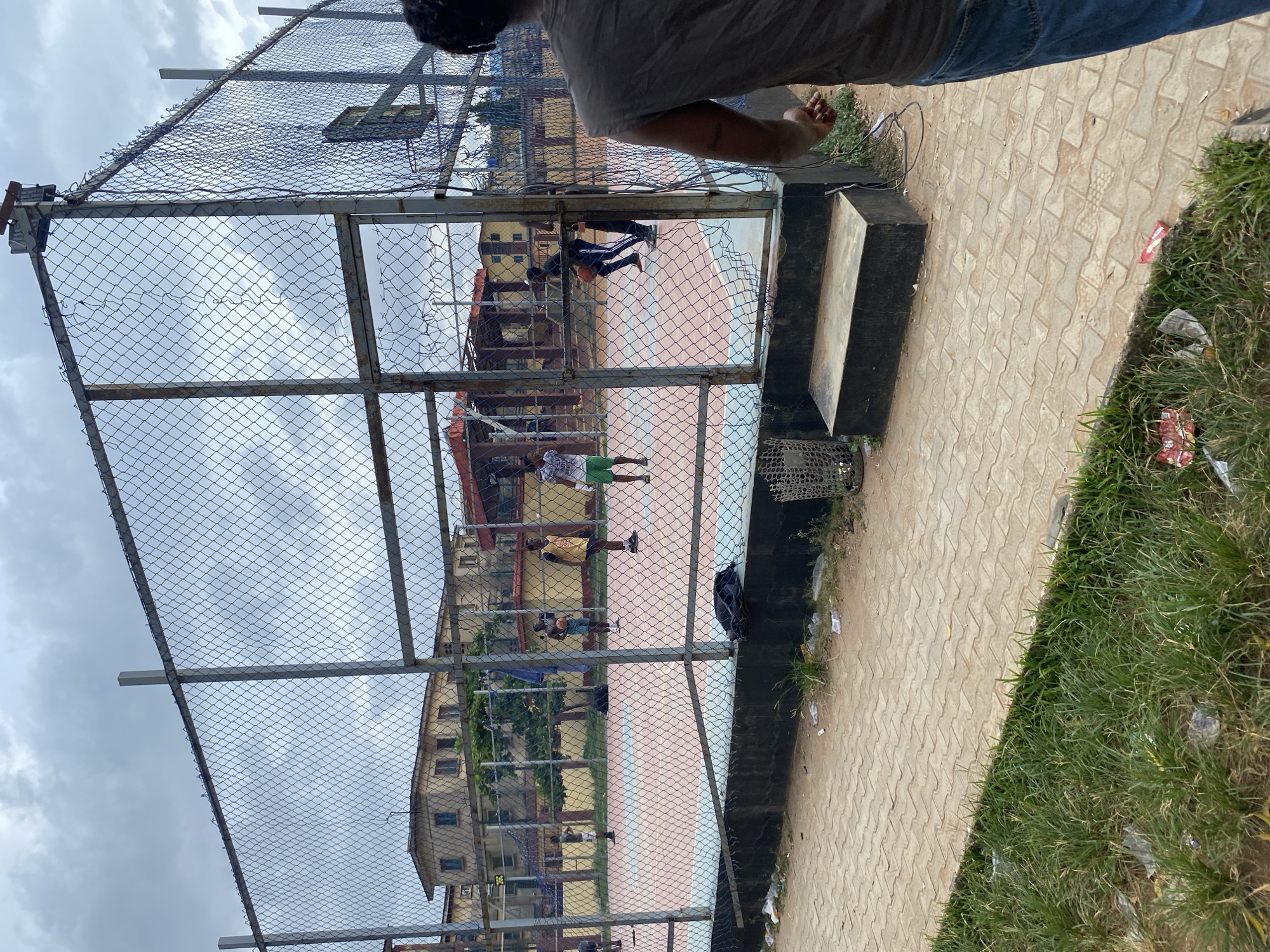 The image captures a view through a chain-link fence, revealing a basketball court where several people are engaged in play. In the immediate foreground, at the top of the frame, the upper body of a person is visible, wearing a grey t-shirt and blue jeans, seemingly looking down into the court.
The fence itself is made of metal mesh and frames the scene, with some visible wear and tear. Beyond the fence, a basketball court with distinct light blue and pink/orange sections is visible. On the court, at least five individuals are present, appearing to be playing basketball. One person wears a white and green outfit, another a yellow shirt, and a third a blue top, with a basketball visible near them.
In the background, behind the court, several multi-story buildings with numerous windows are visible, suggesting a residential or institutional setting. The sky above is overcast with clouds.
The ground outside the fence features a paved area with interlocking concrete blocks, bordered by patches of grass. There is some litter scattered on the grass and pavement, including plastic wrappers and other debris. A dark concrete step or platform is also visible on the right side of the foreground.
The overall scene depicts an active daytime setting, likely a community or school court, under a cloudy sky in an urban environment like Lagos, Nigeria. There is no visible text.
