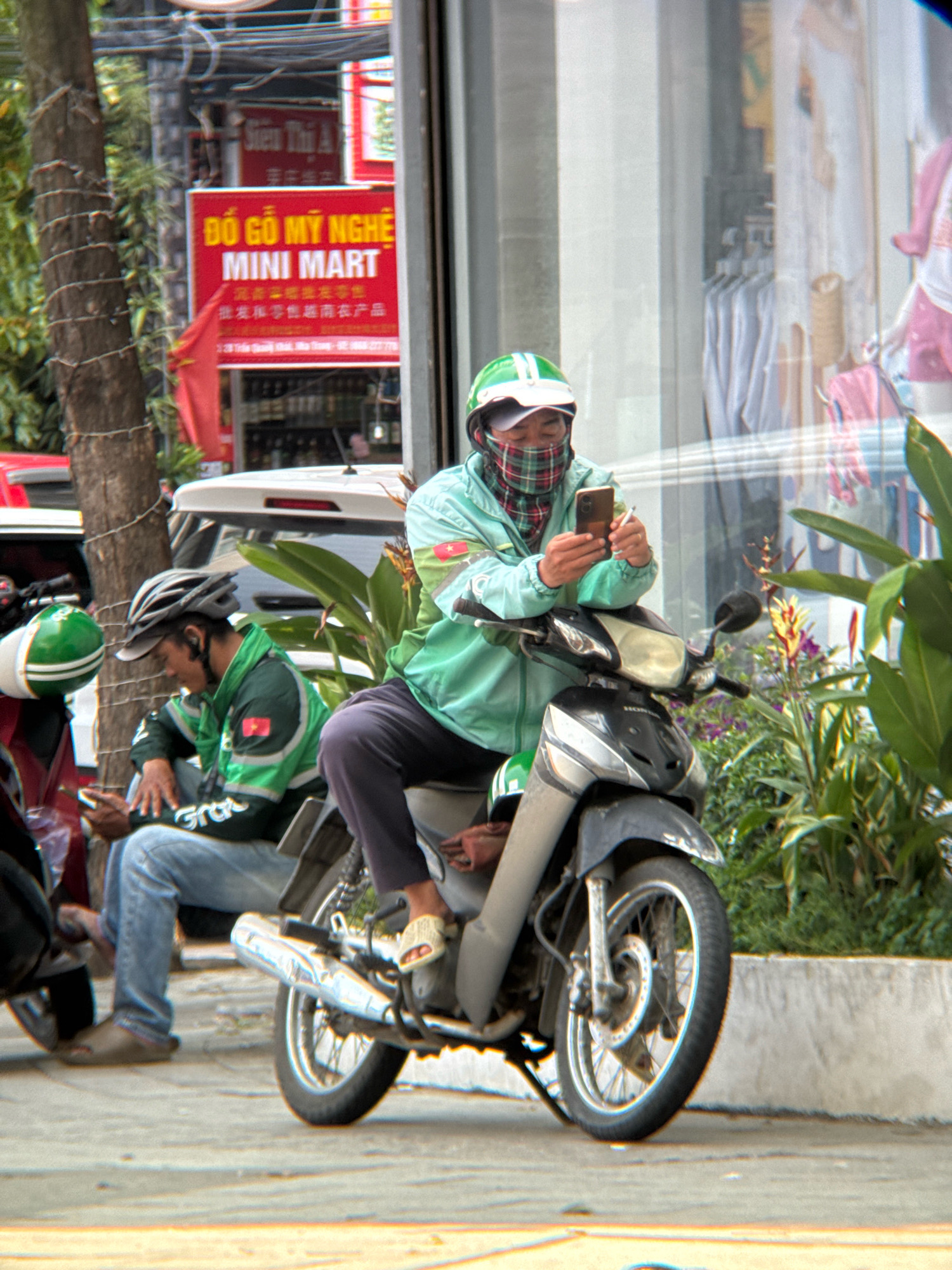 Two men on motorcycles are parked on a sidewalk in Nha Trang, Vietnam. The man on the right wears a green jacket, a checkered scarf covering his face, and a green helmet. He is holding a smartphone and appears to be looking at it. The man on the left wears a green jacket with "Grab" printed on it, a grey helmet, and blue jeans. He is also looking at his phone and has a cigarette in his right hand. A red sign for "MINI MART" is visible in the background. The scene is outdoors, with a building and vegetation visible behind the men.