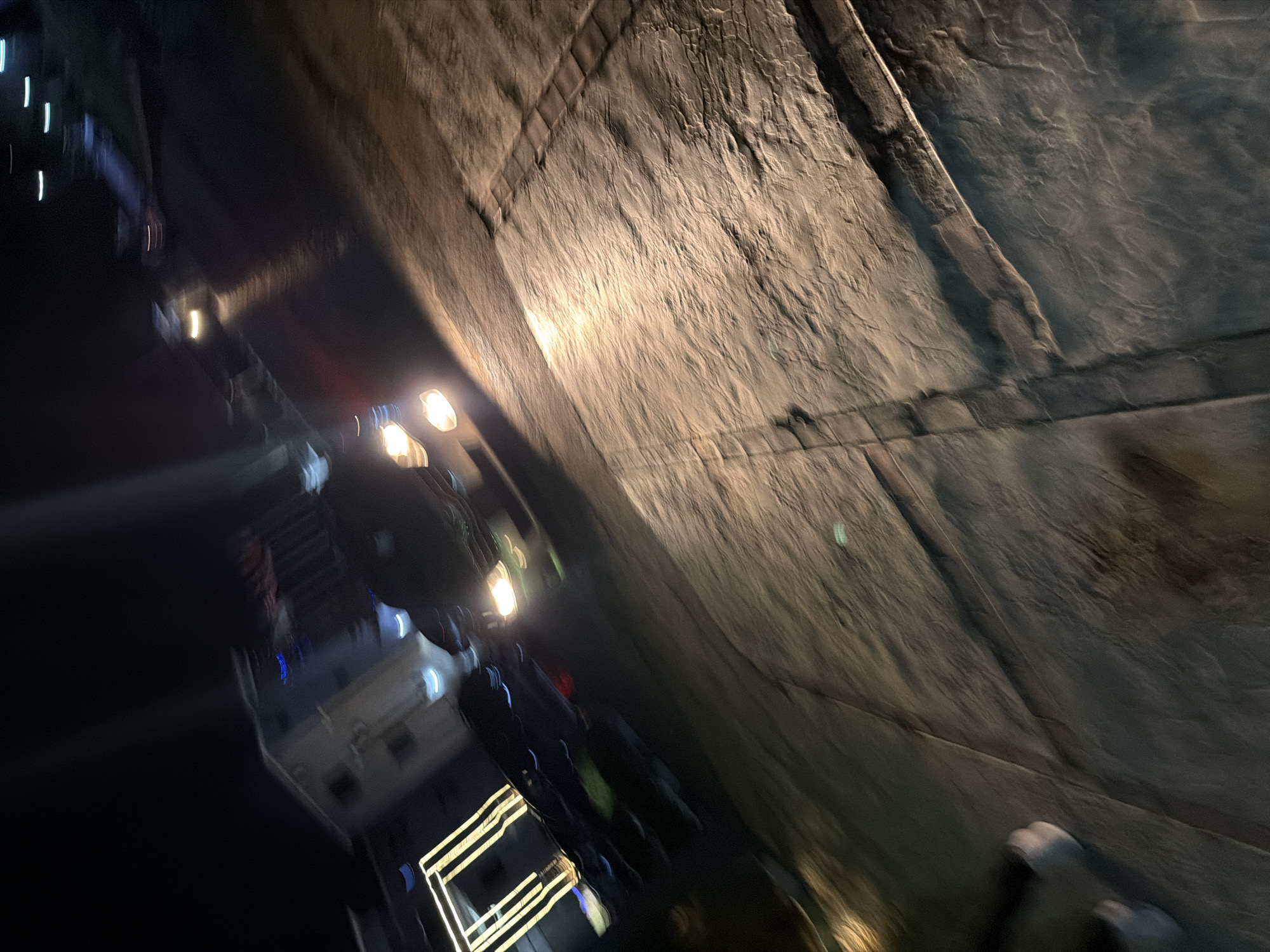 The image captures a night scene on a textured paved surface, possibly a driveway or walkway, with a slight tilt to the right. A dark-colored sedan is partially visible in motion, its headlights casting a bright, blurred illumination on the ground. To the left, a building with distinct rectangular illuminated elements, possibly decorative lighting or windows, is discernible, with a portion of a lighter colored structure also present. The background is dark and indistinct, with streaks of light suggesting other moving vehicles or distant streetlights. The overall impression is one of movement and low-light conditions. While the image depicts a general nighttime urban environment, it does not contain specific landmarks or indicators to confirm its location in Bukuru, Nigeria.