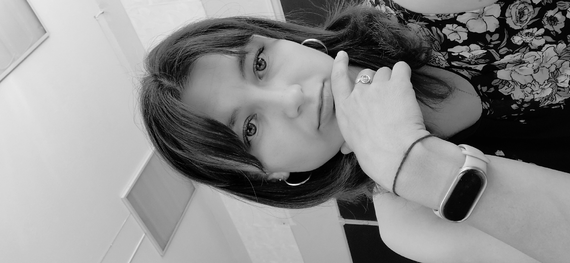 A black and white, medium shot, selfie of a young woman with dark hair and bangs, looking directly at the camera. She is wearing a sleeveless top with a floral pattern. Her right hand is resting on her chin, and she is wearing a ring with a circular stone. She has a black bracelet on her left wrist, and a white fitness tracker. The background is out of focus and appears to be an indoor setting, possibly an office, with a white ceiling and a light fixture. There is no visible indication of the location being Svetlogorsk, Russia.
