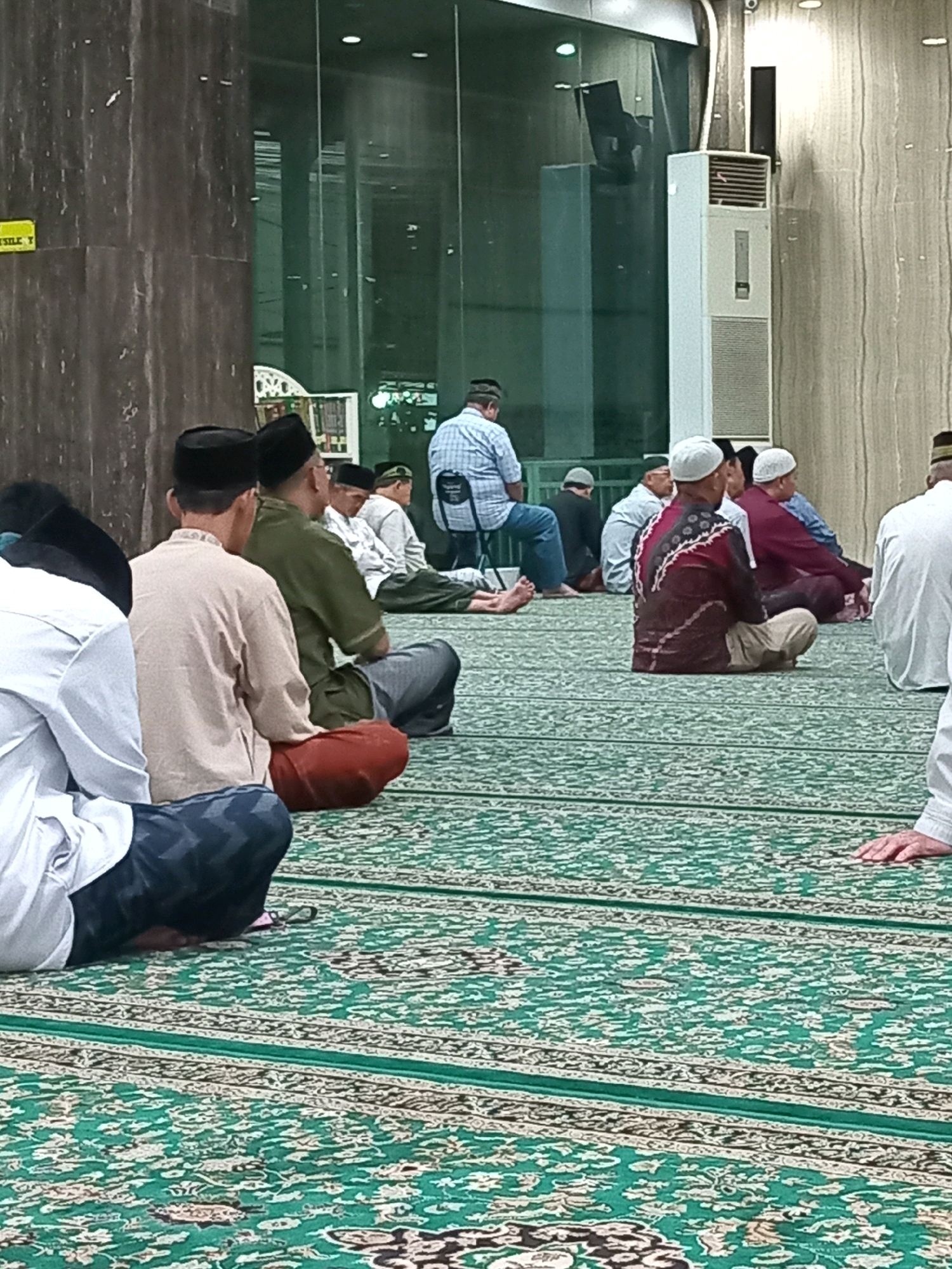The image depicts a group of Muslim men sitting on a green carpet in what appears to be a prayer hall. The men are dressed in traditional attire, including kufis (skullcaps) and sarongs. Some are facing away from the camera, while others are partially visible from the side. The environment includes a large air conditioning unit, glass walls, and tiled surfaces. The carpet features an intricate green and white pattern. The setting is identified as the city of Randuagung, Indonesia.
