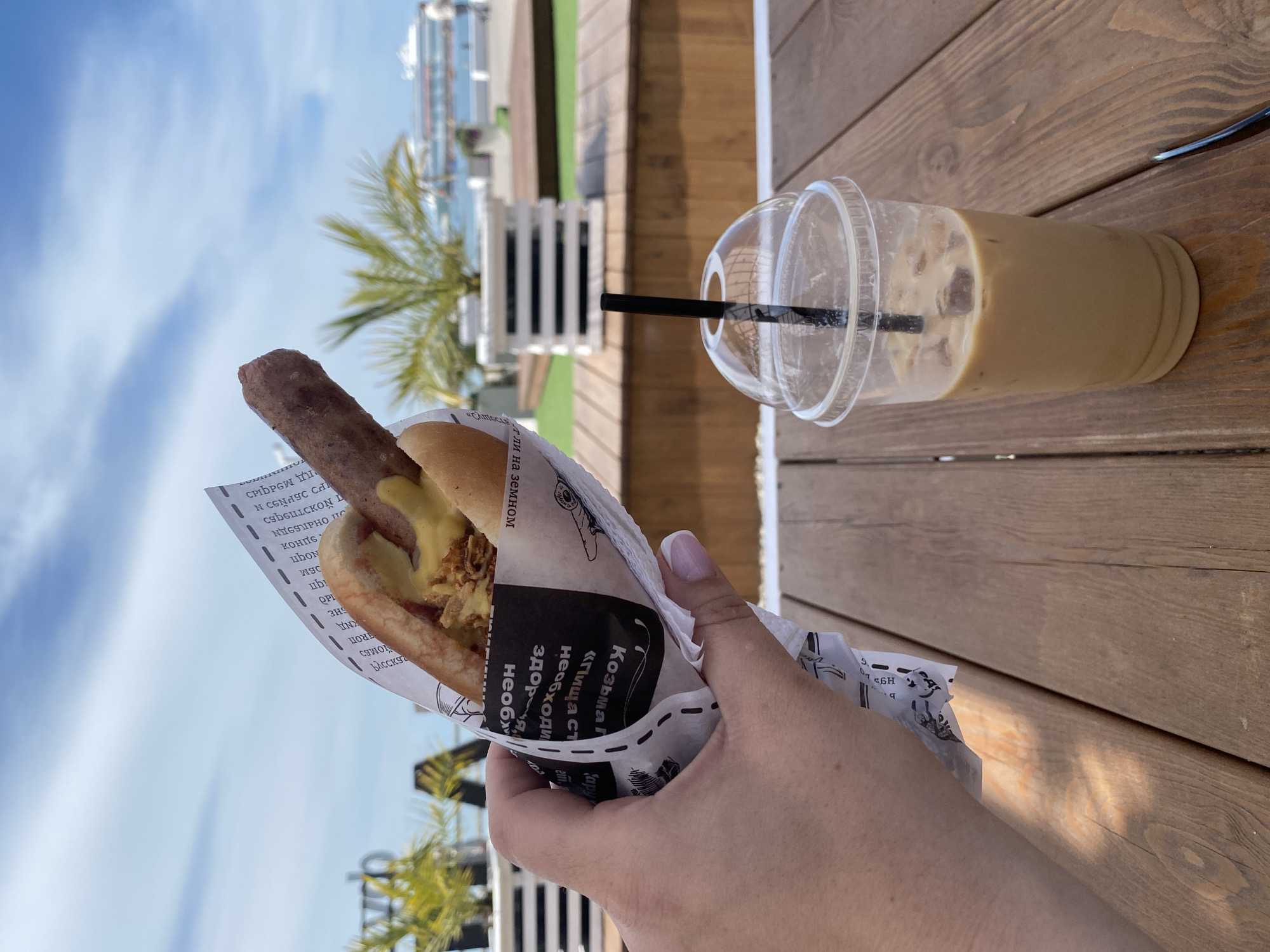 The image shows a hot dog held in a white paper wrapper with Russian text, and a cup of iced coffee with a black straw, resting on a wooden table. The hot dog appears to be topped with mustard and fried onions. A person's hand is visible holding the hot dog. In the background, there is a partial view of what appears to be a beachside area. White planters are visible along with some palm trees. The sky is blue with some clouds. The scene appears to be in Sochi, Russia, based on the Russian text in the hot dog wrapper.