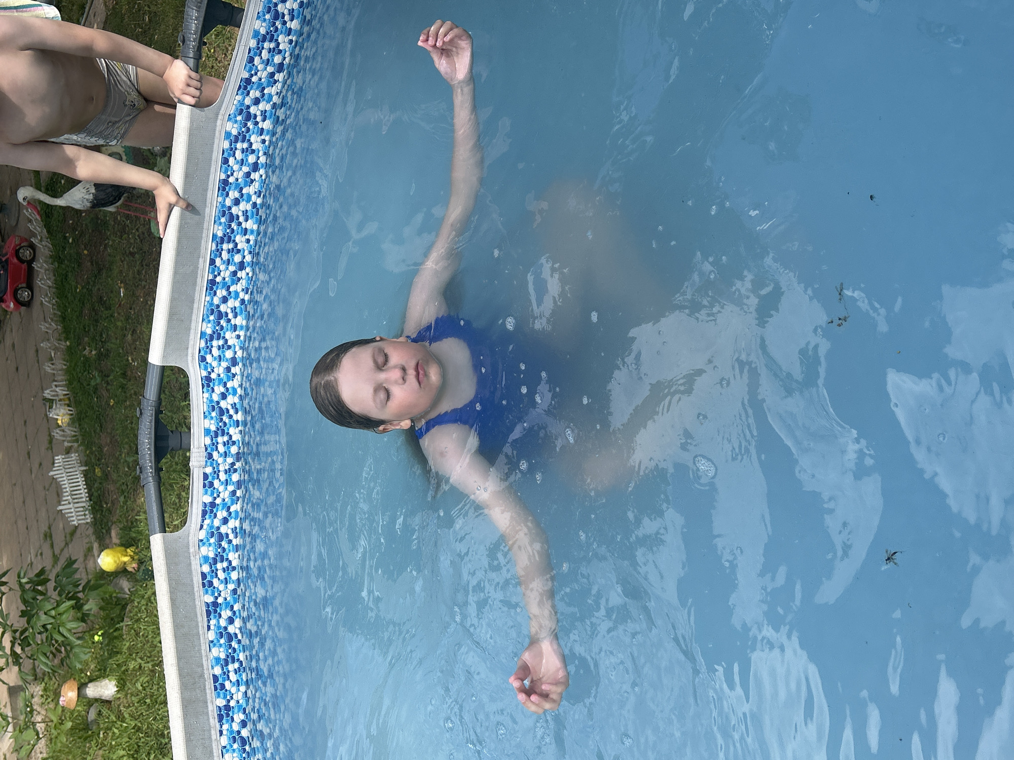The image shows a girl floating on her back in a blue swimming pool with a decorative blue and white tiled border. She is wearing a blue swimsuit. Her eyes are closed and her arms are extended outwards, appearing relaxed. The water is calm with some ripples and small bubbles visible.
In the background, to the left of the pool, there is a person in swim trunks, partially visible and holding onto the pool's edge. Further back, there is grass, a paved area, a toy red car, a decorative stork figurine, and some green foliage.
The scene suggests a warm, sunny day suitable for swimming, likely during daylight hours. The overall atmosphere is one of leisure and enjoyment of a summer activity. There is no visible text in the image.
