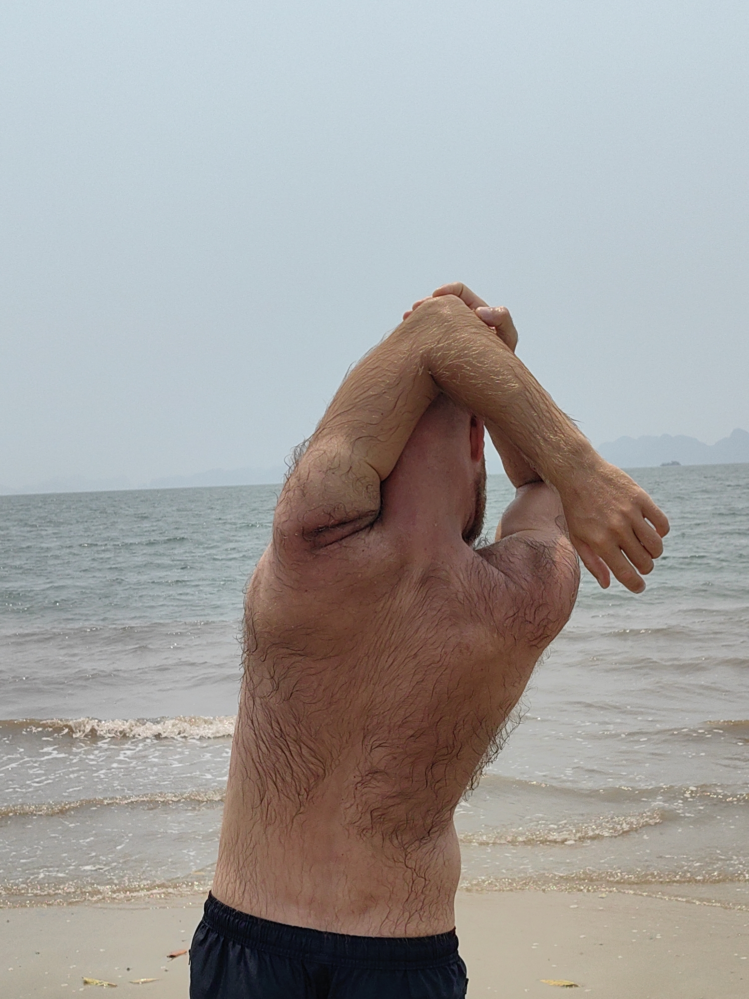 A shirtless man with a hairy back and arms is stretching his arms behind his head on a beach. He is wearing dark shorts. The ocean is in the background with distant landmasses visible. The location is Bai Chay Ward, Vietnam, as suggested by the presence of the sea and distant land features typically seen in that area.