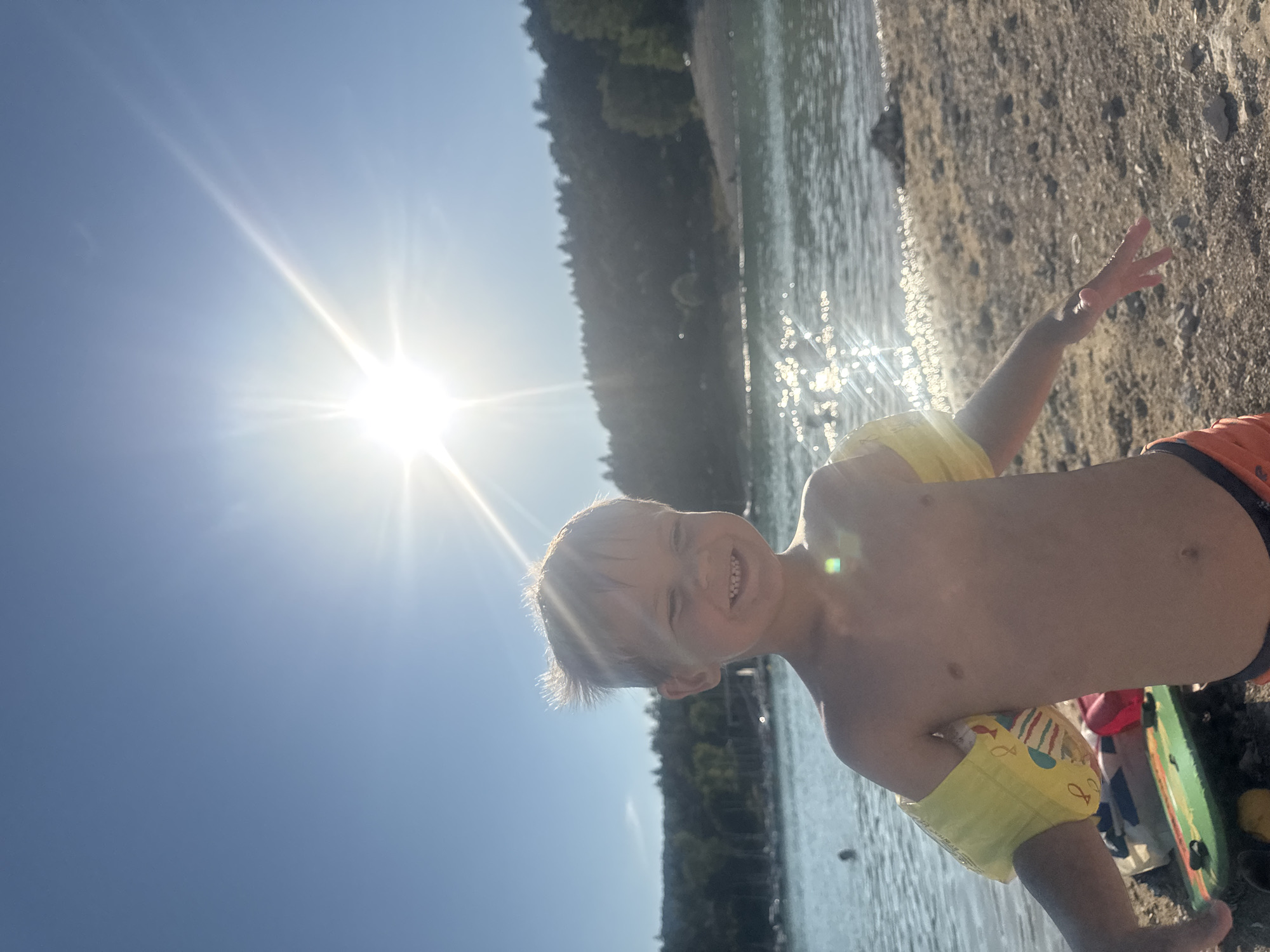A young boy with blond hair and arm floaties is standing on a beach, smiling at the camera. He is wearing orange swim trunks. In the background, there is a body of water, trees, and a clear blue sky with the sun shining brightly. The scene appears to be a lake in the city of Maisod, France.