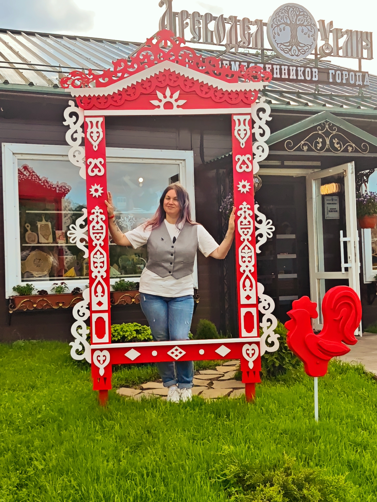 A medium shot depicts a woman standing in front of a decorated red and white wooden frame, located in Chelganovo, Russia. The woman has shoulder-length hair with streaks of purple and is wearing a white T-shirt, a gray vest, and blue jeans. She is standing inside the frame, her hands resting on the sides.
Behind the frame is a brown building with a sign above the entrance written in Russian, with the words "РЕМЕСЕЛ УГЛИЧ" and "РЕМЕСЛЕННИКОВ ГОРОДА", as well as an emblem featuring a tree. The building has a white-framed window display on the left, with wooden items on display. To the right, there is an open door leading inside.
In the foreground, green grass covers the ground, and there is a bright red rooster on a white stick, standing to the right of the frame. The overall scene is well-lit, with the colors appearing bright and vibrant.