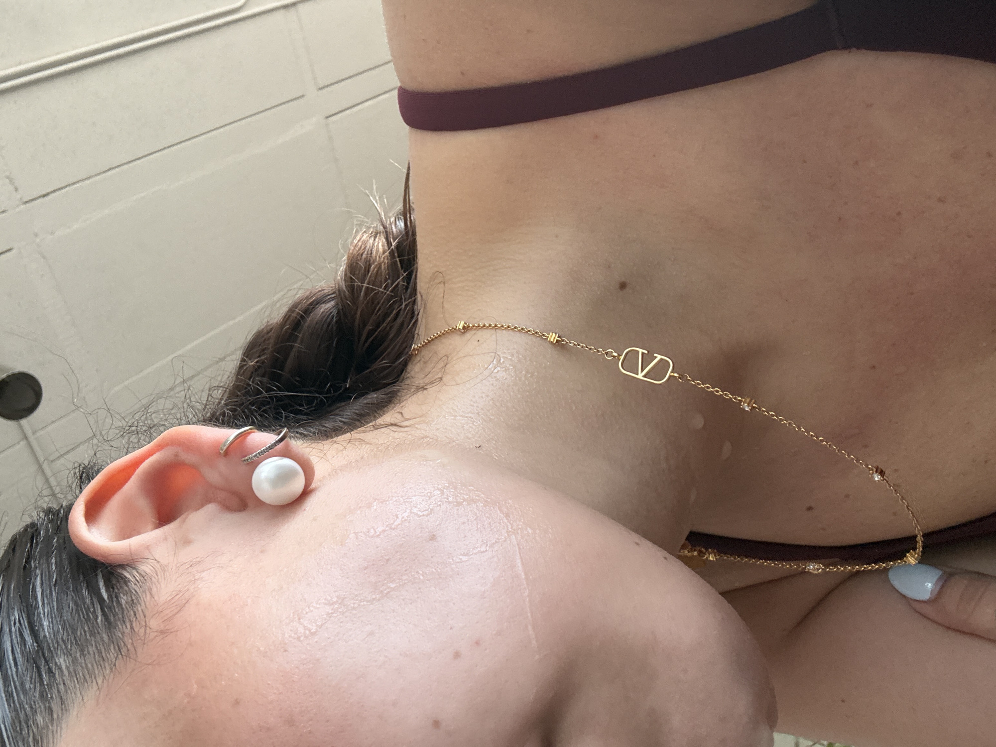 A close-up shot focuses on a person's ear, neck, and upper chest area. The ear has a pearl earring and another ring-like earring. The person has wet hair and dewy skin. The neck is adorned with a gold necklace featuring a "V" pendant, and a burgundy strap is visible, likely a part of clothing. The background is blurry and appears to be a tiled surface. It is not possible to determine if this image was taken in the city of Puebla, Mexico.