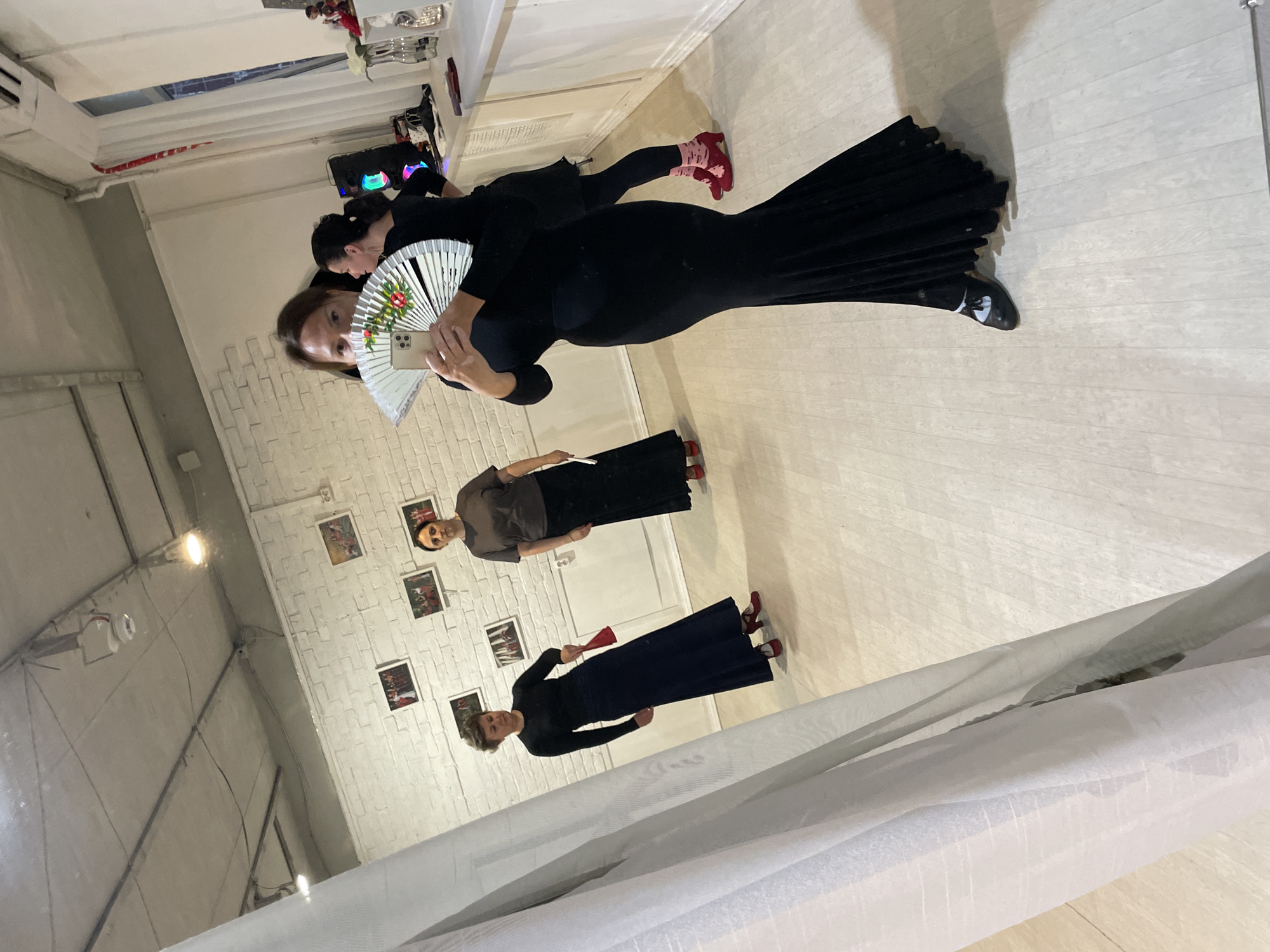 The image, oriented sideways, captures a reflection of four women in a large mirror within what appears to be a dance studio or practice space in Sochi, Russia. The subjects would be standing upright if the image were rotated correctly.
In the foreground, a woman wearing a black long-sleeved top and a long black skirt is taking a selfie, holding a smartphone in her right hand and a large white folding fan with a red and green floral pattern in her left. Her face is clearly visible in the reflection. Slightly behind her, another woman, also dressed in black, is partially visible, wearing pink/red ankle socks and red shoes. She stands near a shelf or table in the background where multi-colored illuminated speakers are placed.
Further within the reflection, two more women are present. One is seen wearing a dark brown or grey top and a long dark skirt, paired with red shoes. The other is dressed in a black long-sleeved top and a long dark skirt, holding a small red folding fan, also wearing red shoes. Their coordinated attire, including the long skirts and fans, suggests they are involved in a dance or performance art, possibly flamenco.
The room features a light-colored wooden or laminate floor and white walls. One wall displays a subtle brick-like texture with several framed pictures hanging on it. Light fixtures are visible on the ceiling, and white sheer curtains or fabric are draped in the lower right portion of the image. The scene conveys a moment of practice or preparation during a group session. There is no visible text.