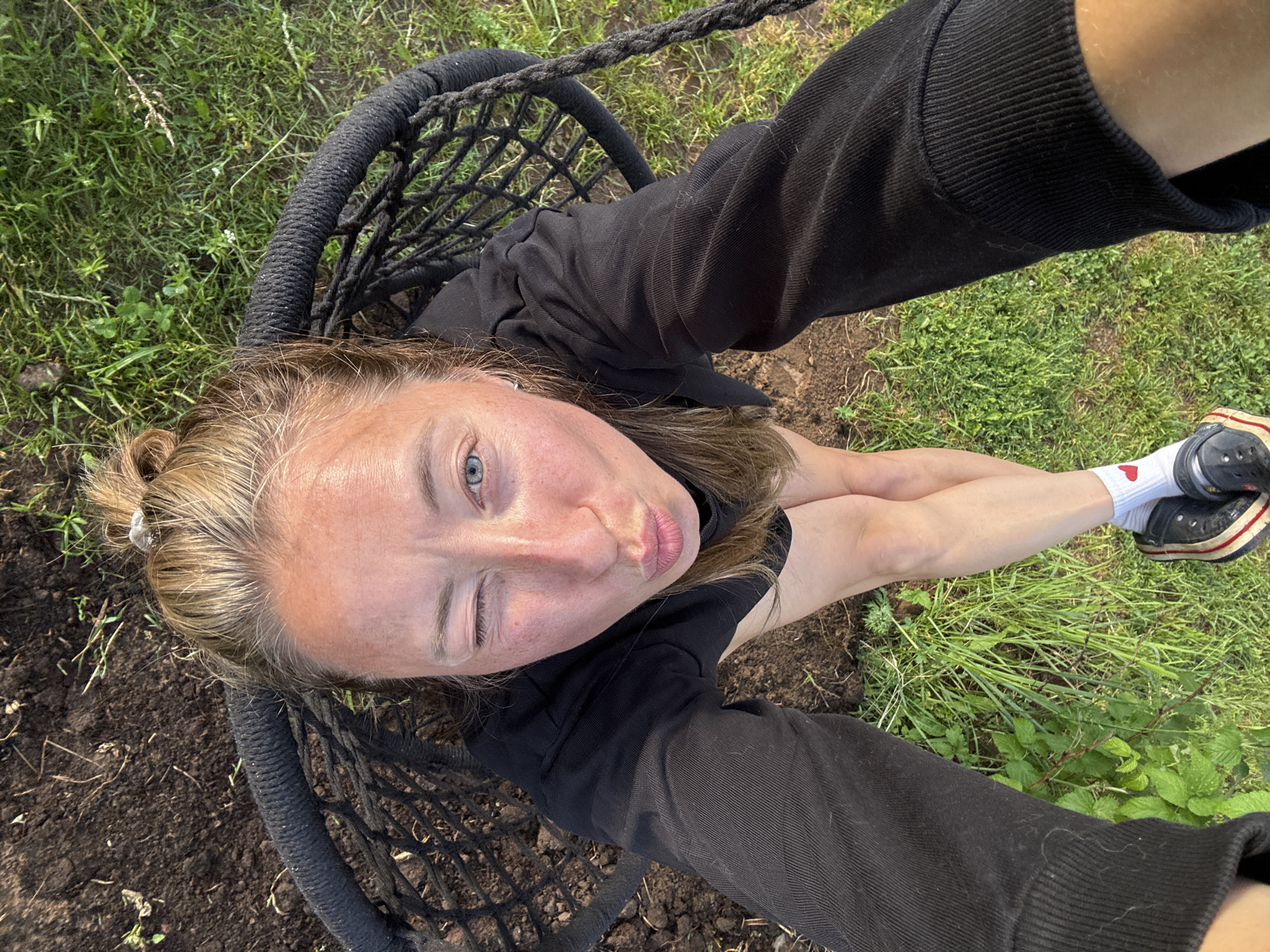 A young woman with blonde hair pulled into a bun is lying down, taking a selfie. She is wearing a black sweatshirt and black pants. Her legs are crossed and she is wearing white socks with a small red heart on them and black and red striped shoes. The scene is outdoors with grass and dirt. The woman has a playful expression, with her eyes closed and her lips pursed. The lighting suggests it is daytime. The background shows green grass and some dirt.
