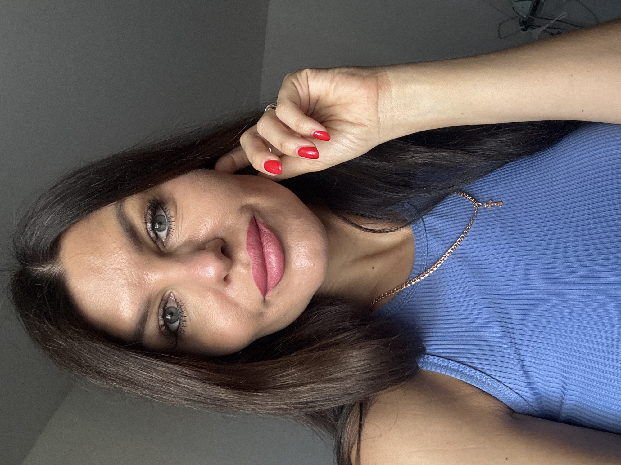 The image shows a close-up, head-and-shoulders portrait of a woman with dark brown hair, light green eyes, and pink lipstick. She is wearing a blue, ribbed tank top and a rose gold chain necklace with a small cross pendant. Her right hand is raised, touching her right ear. Her fingernails are painted bright red. The background is a plain, dark gray wall. The image is taken indoors with soft lighting. The location in Chaikovsky, Russia is not discernible from the visual information provided in the image.
