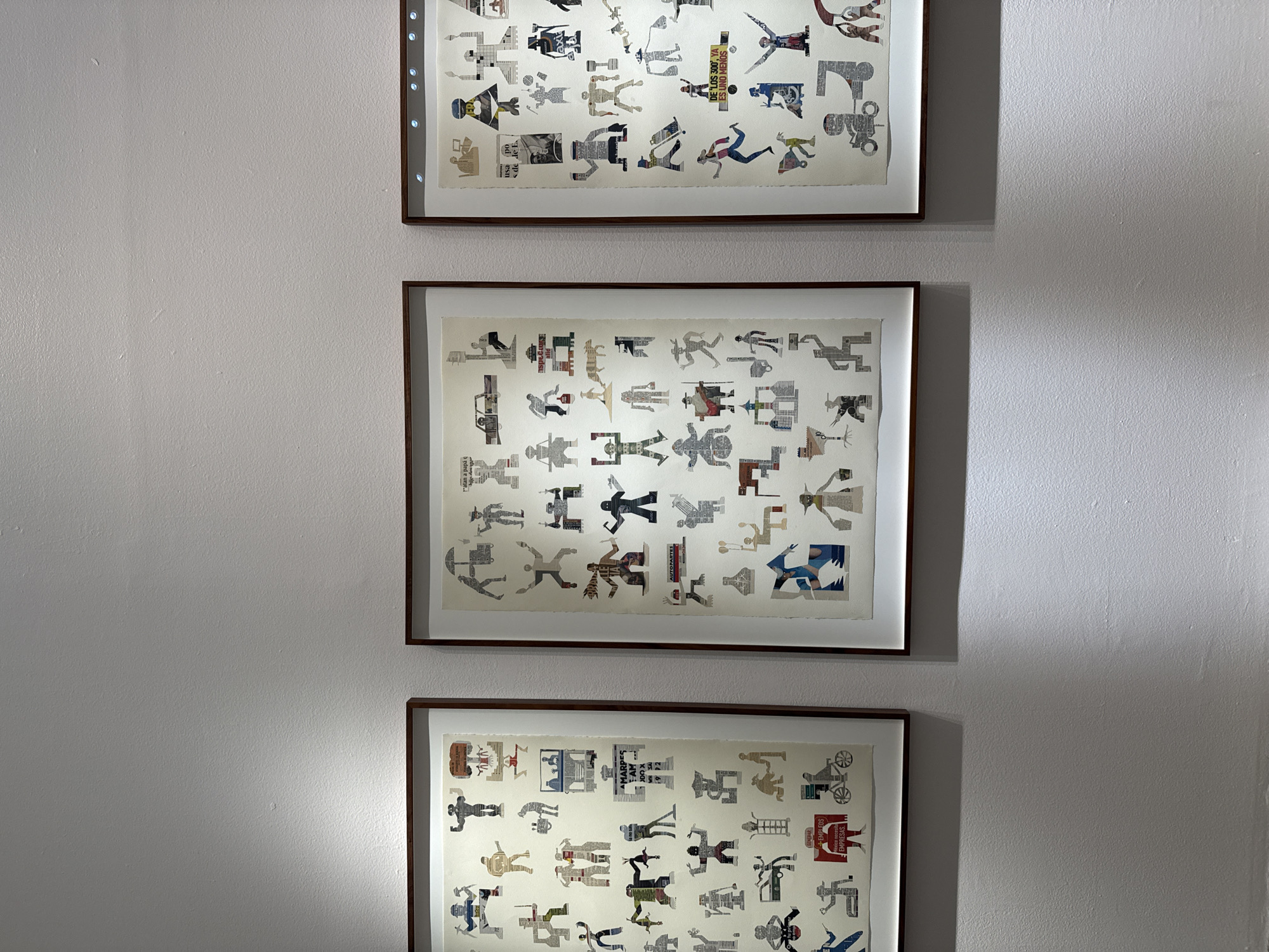 The image shows a vertical arrangement of three framed pieces of art hanging on a plain, likely off-white, wall. Each piece appears to contain numerous figures cut from printed material like newspapers or magazines, arranged in rows and columns. The figures vary in shape and size, depicting human-like forms in different poses. The frames have a narrow, dark-colored border. The top frame is lit with a light emitting diode (LED) strip along the top. The lighting creates some shadows on the wall and on the frames themselves. The image appears to be taken in a gallery or a home. Some of the newspaper clipping text that can be read contains the words "GENERAR EMPRESAS," "AMARPES FAM JDO X," "atan a papá q," "usa po de de E," "AUTOPARTES," and "DE 'LOS 300', YA ES UNO MENOS." The reference to "Mexico City, Mexico" is implied by the Spanish text in the clippings.