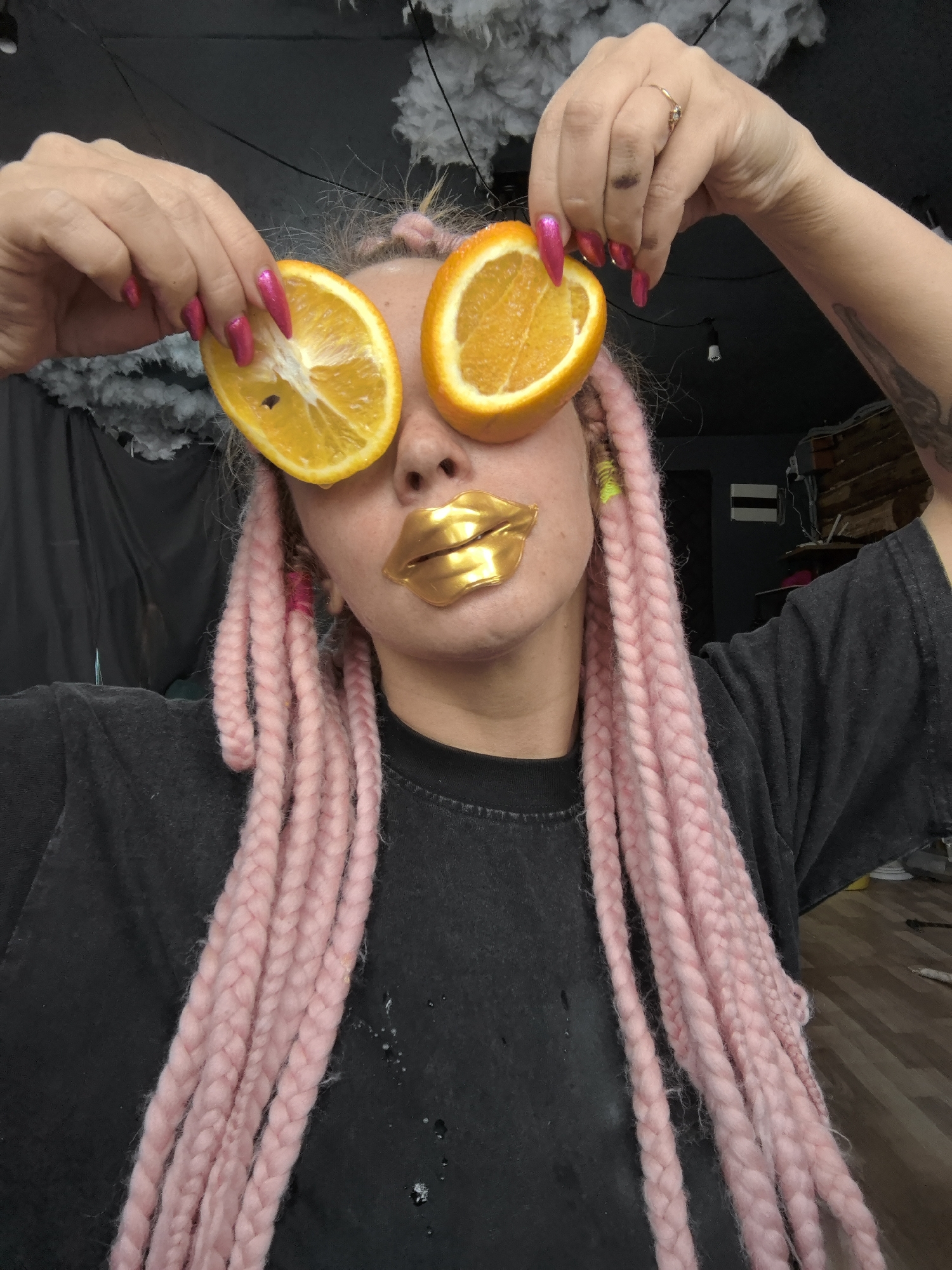 A fair-skinned person with pink braided hair is holding two halves of oranges over their eyes. The person has gold-colored lip coverings. They are wearing a dark gray T-shirt. Their fingernails are painted with a hot pink color. The background appears to be a dimly lit room with various objects and materials. There appears to be what looks like dark colored curtain behind them.
The location cannot be determined from the given image.