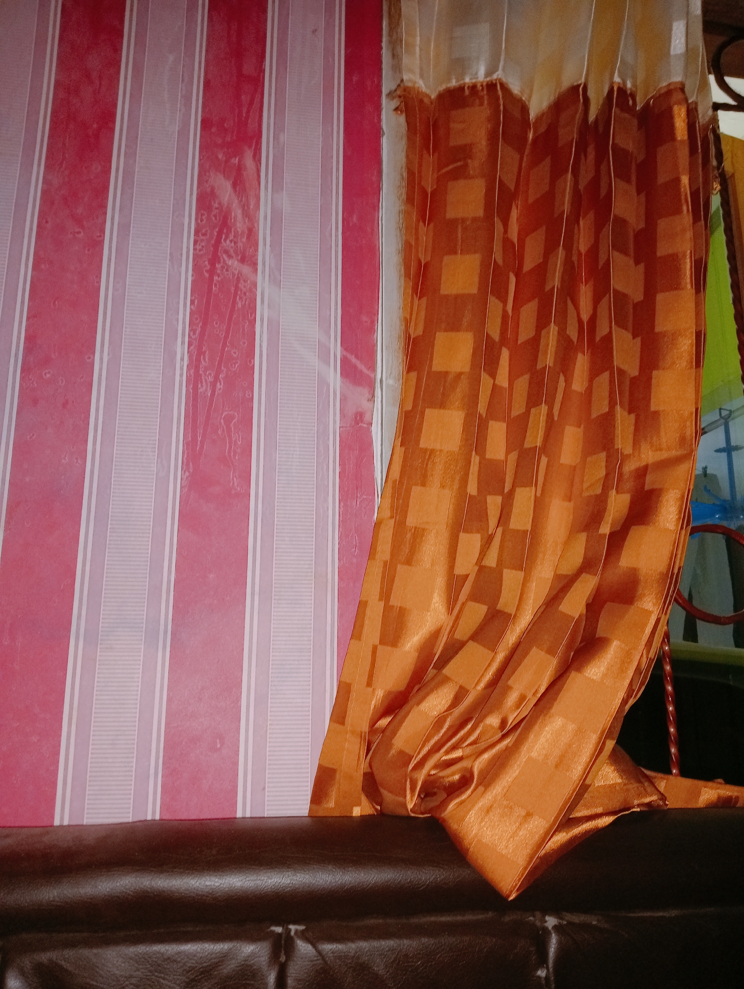 The image depicts a close-up view of interior furnishings. On the left side, a wall is covered with pink and white striped wallpaper. To the right, an orange curtain with a subtle checkered pattern hangs, partially obscuring the wallpaper. The curtain is draped over the back of a dark brown leather sofa, visible in the foreground. The lighting suggests an indoor setting, possibly a living room or bedroom. There are no people or identifiable landmarks indicating a specific location within Tridayasakti, Indonesia, or anywhere else.