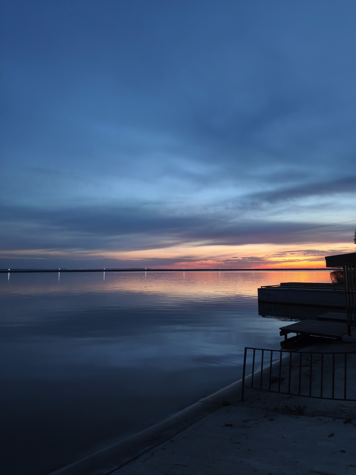 The image depicts a tranquil scene in Chaikovsky, Russia, at dusk. A calm body of water stretches towards the horizon, reflecting the gradient of the sky above. The sky transitions from a deep blue at the top to a vibrant orange and pink near the horizon. There are what appear to be lights on the far bank of the water.
A dark structure with multiple levels, possibly a deck or platform, juts out into the water on the right side of the image. A metal railing runs along the edge of a concrete area in the foreground, providing a sense of enclosure. The overall atmosphere is peaceful and serene.