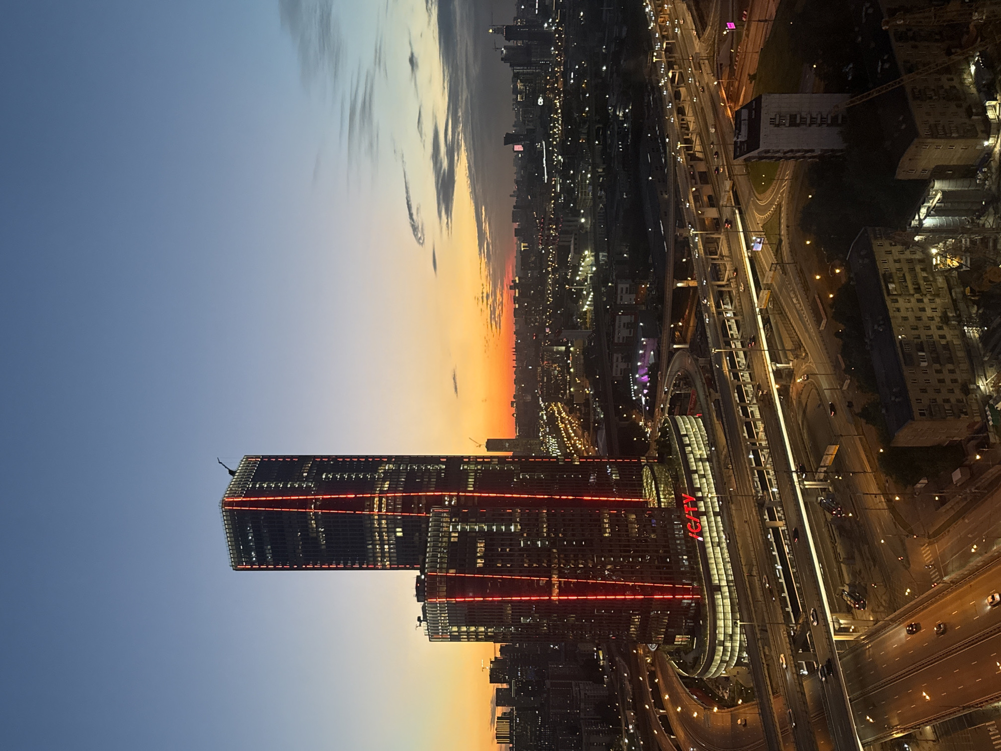 This is an aerial, wide-angle, nighttime photograph of the Moscow City skyline in Khimki, Russia, taken during twilight. The sky transitions from a deep blue at the top to a soft orange and pink gradient near the horizon, with scattered clouds. The focal point of the image is a modern skyscraper with a distinctive red lighting design down its facade, illuminating vertical stripes on the dark glass. Adjacent to it is another building of similar architectural style, also lit with red accents. The base of the main skyscraper features a circular structure with the word "ICITY" displayed in red neon letters on its facade.
Surrounding the skyscrapers is a complex network of elevated highways and roads, with visible traffic and streetlights. Cars are visible on the roads, appearing as streaks of light and individual vehicles. Several other buildings, varying in height and architectural style, are spread across the cityscape, many illuminated with lights. In the background, further buildings and structures, some appearing industrial or under construction with cranes, dot the horizon. The overall scene depicts a bustling urban environment at dusk, characterized by modern architecture, extensive infrastructure, and the ambient glow of city lights against the fading natural light. There are no visible people in the image.
