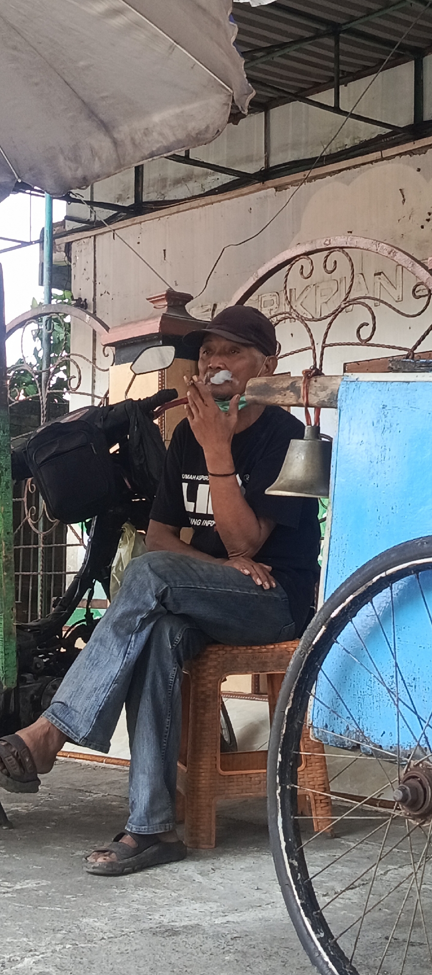 In Krian, Indonesia, a man is seated on a brown plastic stool, smoking a cigarette. He wears a dark baseball cap, a black t-shirt with white lettering, and blue jeans. His right hand holds the cigarette to his mouth, with smoke visible. A bicycle wheel is prominent in the foreground on the right side of the image. Behind the man, a blue cart-like structure is visible, and a bell hangs from a wooden pole attached to it. A black bag is slung over a motorcycle in the background, which is partially obscured by a decorative metal gate. The environment appears to be outdoors, possibly a street or a market, with an awning providing shade.