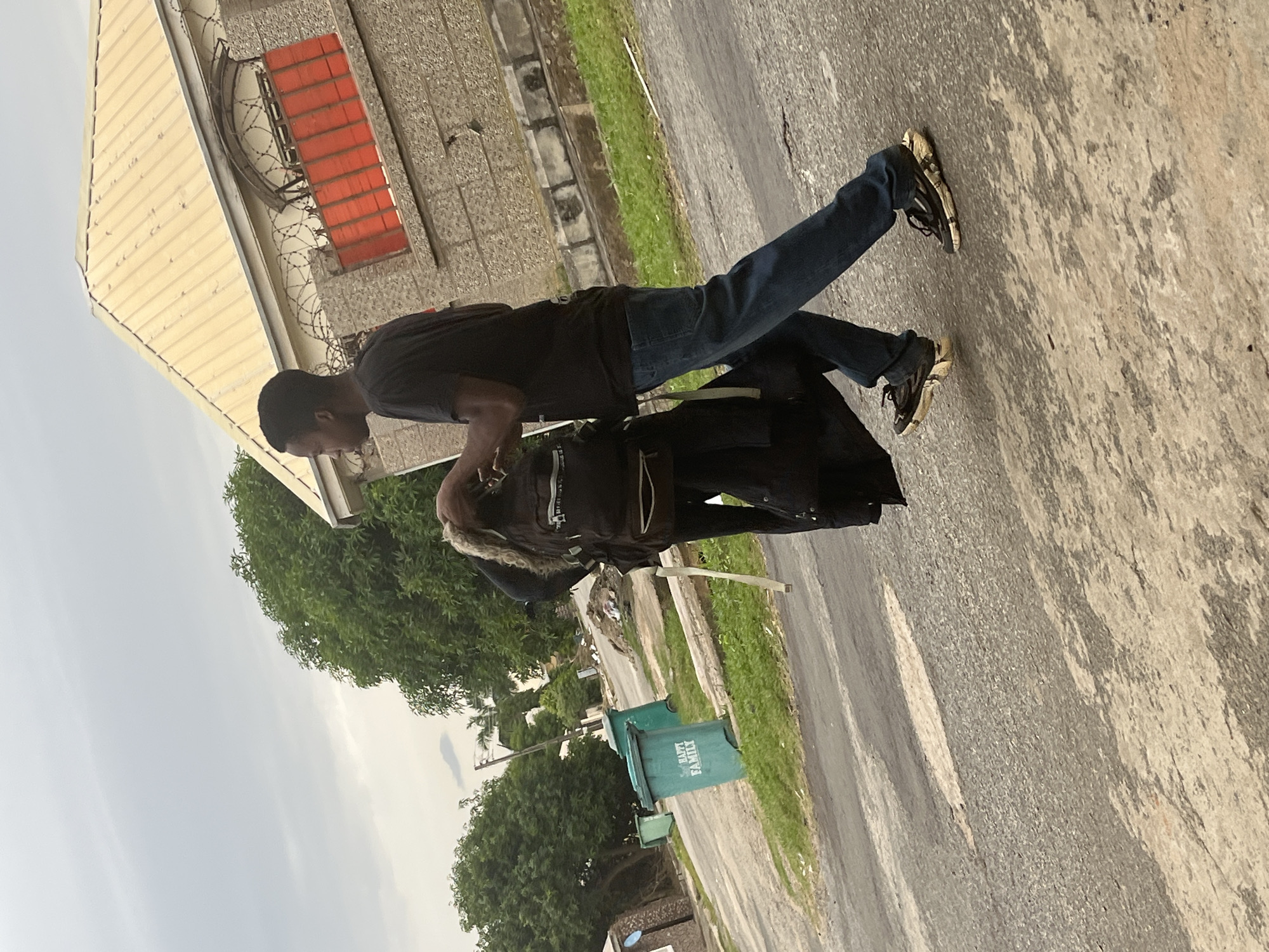 The image depicts a street scene in Abuja, Nigeria, under an overcast sky. In the foreground, a young Black man wearing a black t-shirt and blue jeans walks towards the right, carrying a black backpack over his left shoulder and a folded black garment in his right hand. His right foot is positioned forward, and his left foot is slightly behind. He is wearing black sneakers with white and gold accents. To his left, another person, whose face is not visible, is also seen carrying a backpack. In the background, a beige-colored building with a metallic roof and red window frames is visible, surrounded by a stone wall topped with barbed wire. To the left of the building, several green trash bins are placed along the roadside. Trees with lush green foliage are scattered throughout the scene. The ground is paved with asphalt, showing some wear and tear.