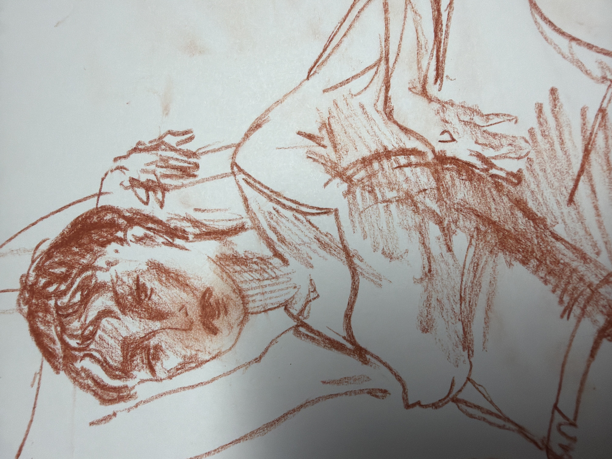 A sketch in reddish-brown crayon on white paper depicts a close-up view of a person from the chest up. The figure is positioned at an angle, with the head slightly tilted to the left. The face is detailed with wavy hair, dark eyes, a nose, and a mouth. The body is adorned with a garment that appears to be a simple dress or tank top, with straps visible over the shoulders. The sketch captures the form of the chest and shoulders, and one arm is bent, with the hand resting near the face. The background is plain white, suggesting the sketch was made in an art studio or on a plain surface. This image is not located in Dongducheon-si, South Korea; it is a drawing.