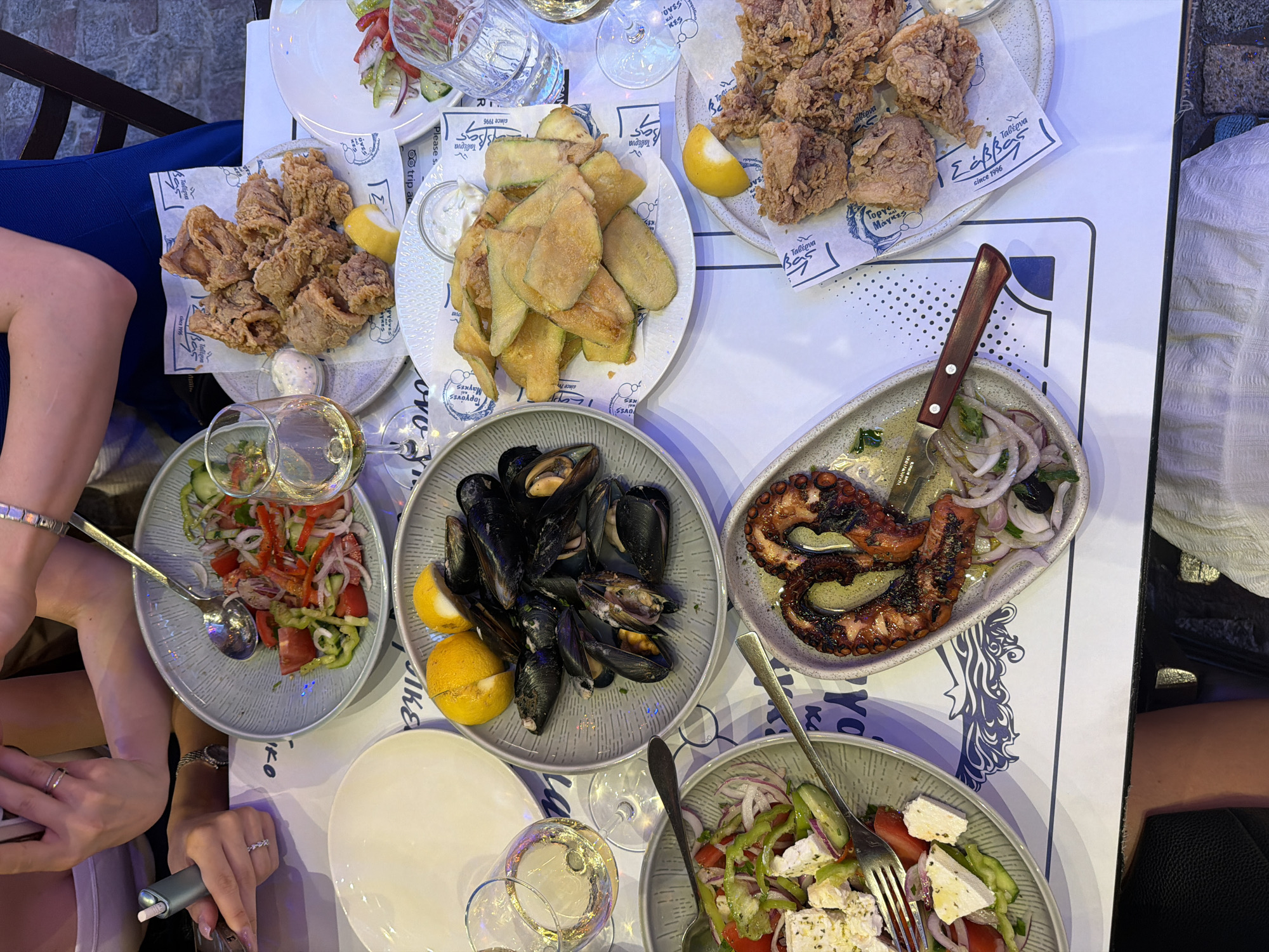 The image depicts a table laden with Greek food, likely at a restaurant in Kavala, Greece. Dishes include fried calamari, fried zucchini, mussels, grilled octopus, and Greek salad. There are also glasses of white wine. Two people are partially visible at the table. The table is covered with a paper tablecloth printed with Greek designs and the name of the restaurant, "Taverna Gorgones Mages". A knife rests on the edge of the octopus dish.