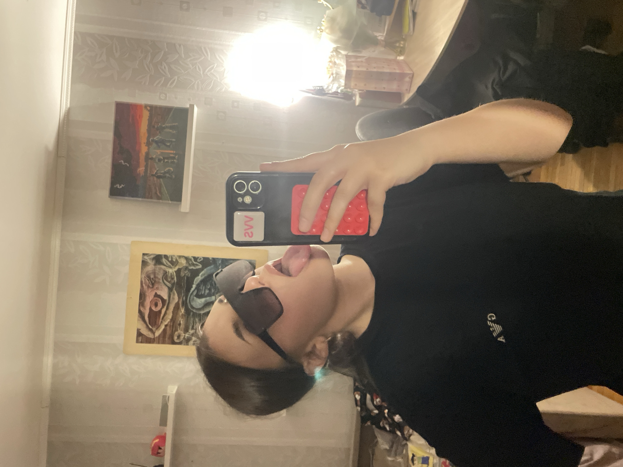 This image is a mirror selfie taken indoors. A young person, likely female, is shown from the chest up, striking a playful pose with their tongue sticking out. They are wearing dark, rectangular sunglasses and a black short-sleeved t-shirt with a small, stylized "GA" logo (Giorgio Armani) visible on the left chest area. Their dark hair is pulled back, partially revealing an ear.
The person is holding a black smartphone with a red pop-it style grip attached to the back. The phone's dual camera lenses are visible, along with a white sticker bearing the red text "2VV" on the lower left of the phone case.
The background, a reflection in the mirror, reveals an indoor setting, possibly a bedroom or living space. The walls are covered with light-colored wallpaper featuring a subtle leaf or floral pattern. Two framed pictures adorn the wall: one depicting a sunset or landscape scene with dark figures, and another appearing to be a more abstract or surreal artwork with organic shapes and colors. A bright, circular light source is prominently reflected in the upper center of the mirror. A wooden floor is visible at the bottom of the reflected scene, and some light-colored furniture or shelving can be seen to the left.
The overall mood is casual and lighthearted, suggested by the playful expression and mirror selfie format. The location context provided is Vsevolozhsk, Russia.