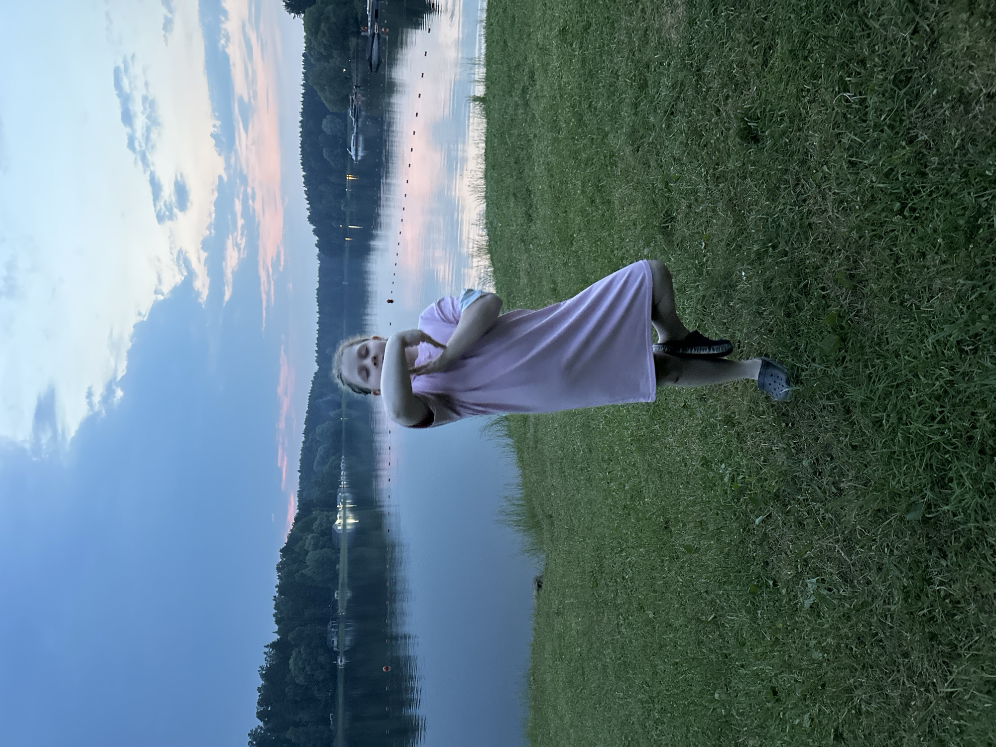 A young girl with blonde hair is lying on a grassy bank next to a large body of water, possibly a lake or river. She is wearing a light pink dress and dark shoes. Her arms are crossed over her chest, and her eyes are closed, suggesting she is relaxed or resting. The water behind her reflects the sky, which is a mix of deep blue and soft pinks and oranges, indicating it is either early morning or late evening. The sun is not visible, but the colors in the sky suggest a sunset or sunrise. Along the far bank of the water, there is a dense line of trees, and scattered lights from what appear to be buildings or houses are visible. There are also some buoys or markers in the water. The overall mood of the image is peaceful and serene. The location is identified as Sokolovo, Russia.