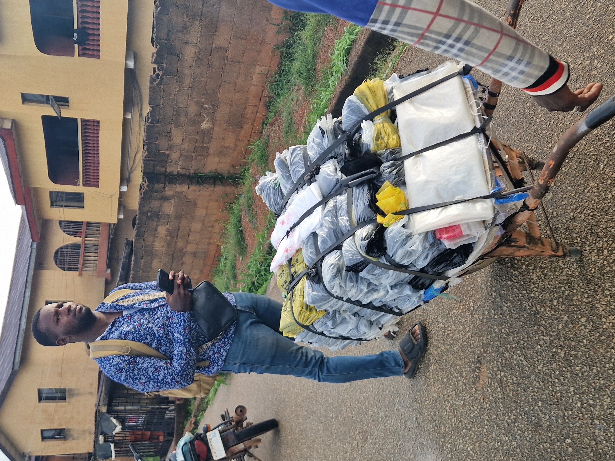 In Kabong, Nigeria, a man stands by a cart filled with goods, holding a mobile phone. He is wearing a blue patterned shirt, blue jeans, and black sandals. A backpack is slung over his shoulder, and a black bag is across his chest. The cart is laden with items wrapped in plastic bags, secured with black straps. Behind him, a building with yellow walls and balconies with red railings is visible. A portion of a red brick wall runs along the side of the street, with green vegetation growing along its base. A motorcycle is parked to the left of the man. Another person, wearing a checkered shirt, is partially visible on the right side of the frame, with their hand near the cart's handles.