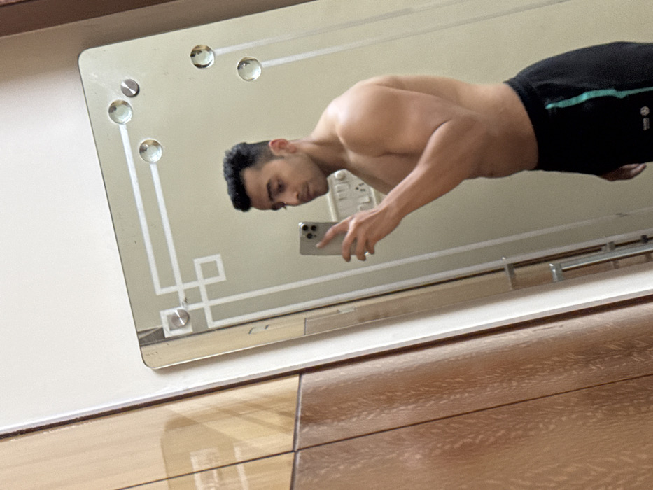A shirtless man in black athletic shorts takes a mirror selfie with an iPhone. He has dark hair and is positioned at an angle, showing off his physique. The mirror is decorated with small, rounded glass accents and a geometric design. The background includes a wall and a dresser with wood grain detail. The image is bright and clear.