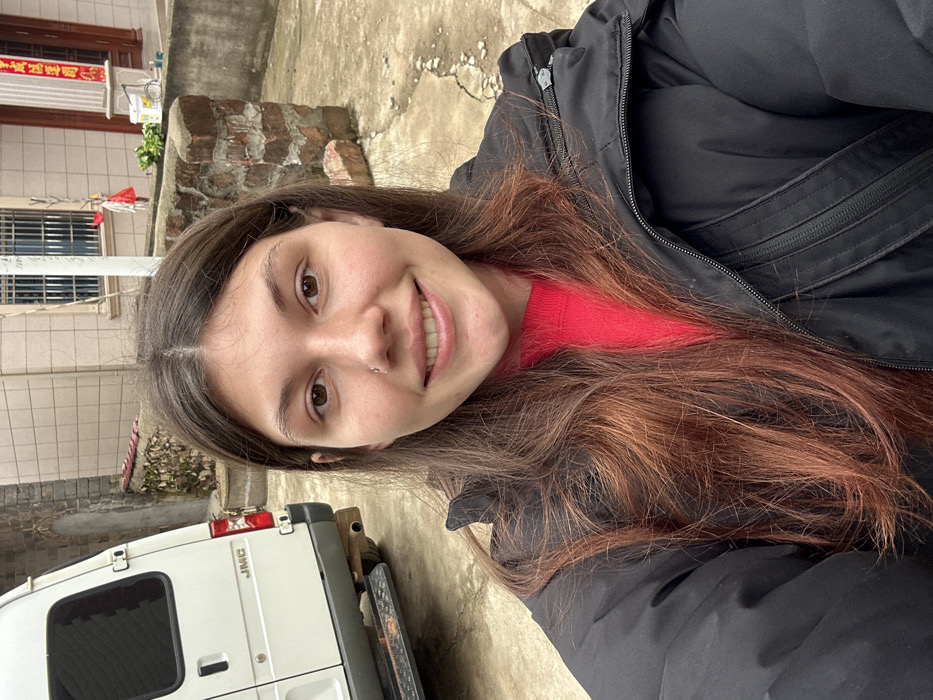 A smiling woman with long, auburn-tinged brown hair takes a selfie outdoors. She wears a red sweater under a black jacket. In the background, a white JMC van sits next to a brick structure and a building with tiled walls and a window with bars. The setting appears to be a narrow alleyway or courtyard. The image is slightly rotated counter-clockwise.
